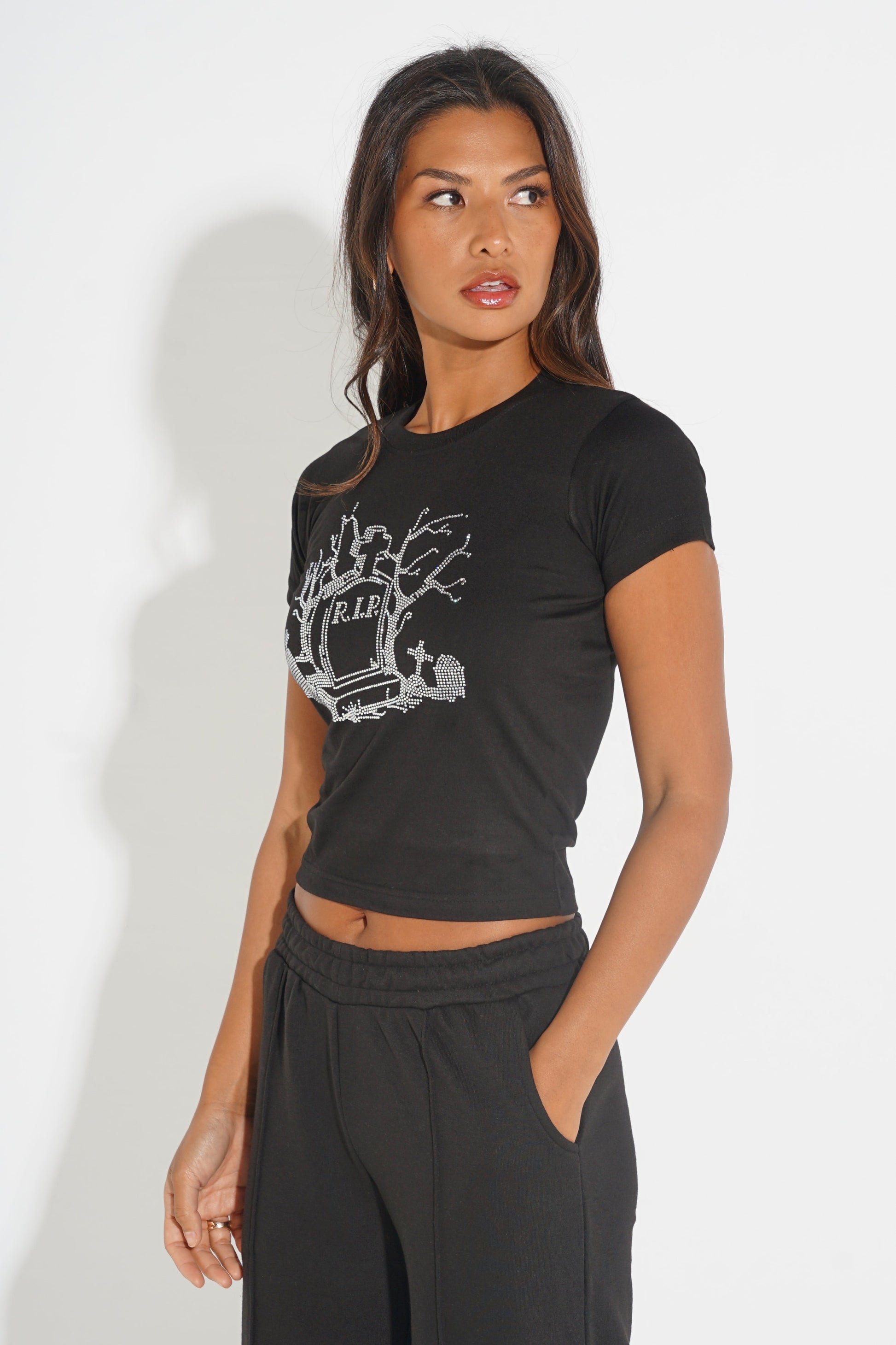 A woman modeling a black short-sleeve crop top with a distressed white graphic print on the front, paired with black sweatpants, against a plain white studio background.