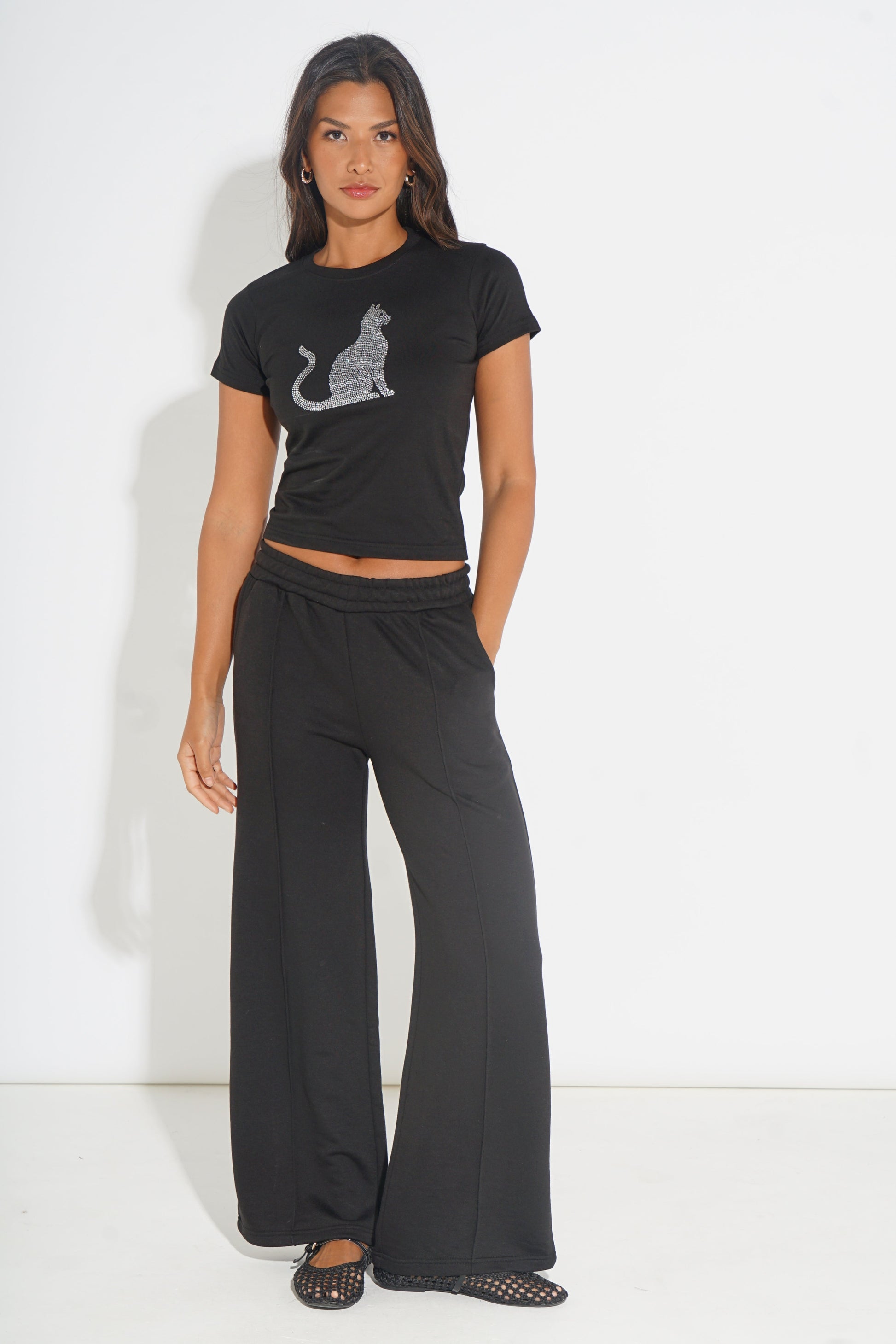 A woman modeling a black short-sleeve crop top with a glittery cat graphic and matching black wide-leg pants against a plain white studio background.
