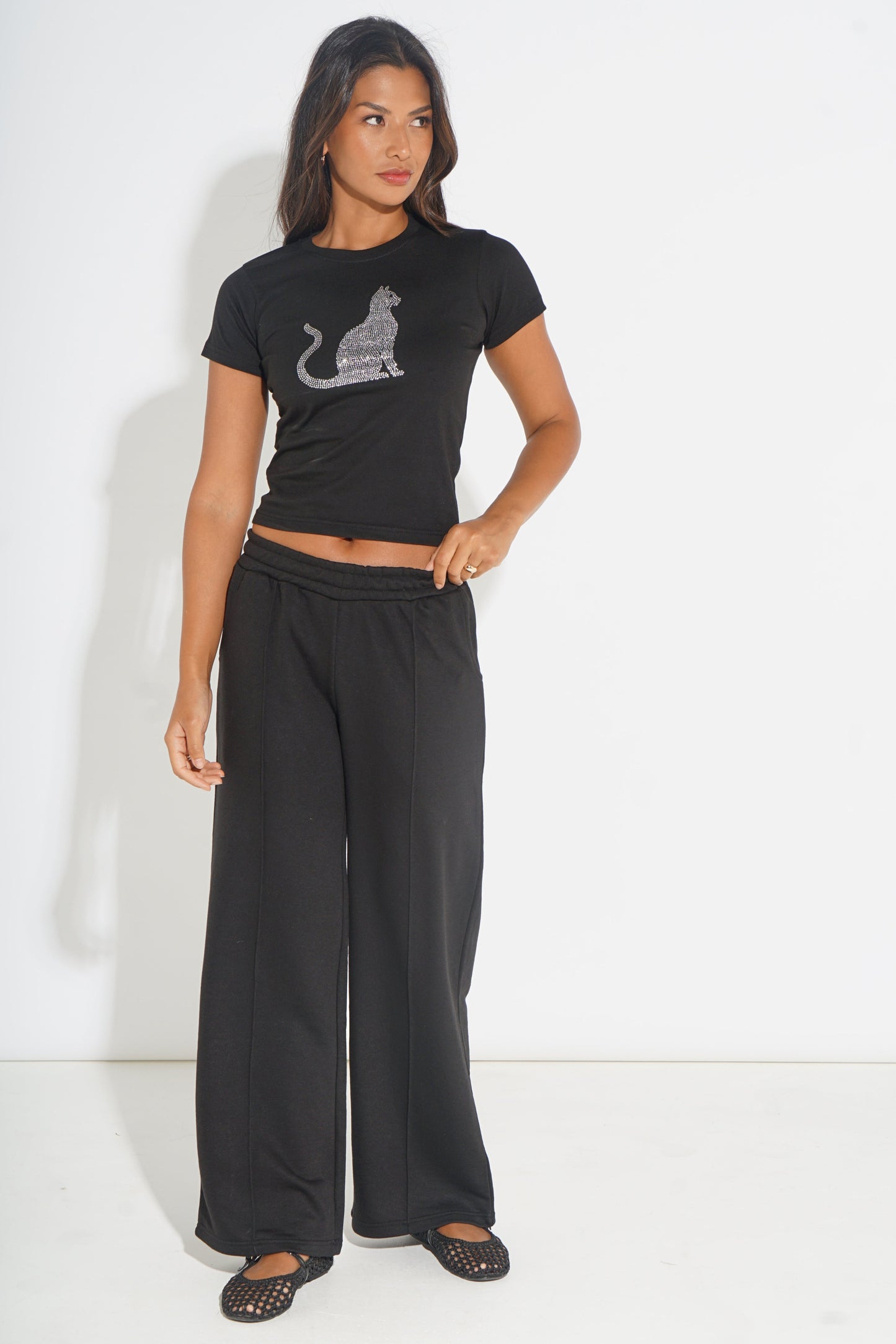 A woman modeling a black short-sleeve crop top with a glittery cat graphic and matching black wide-leg pants against a plain white studio background.