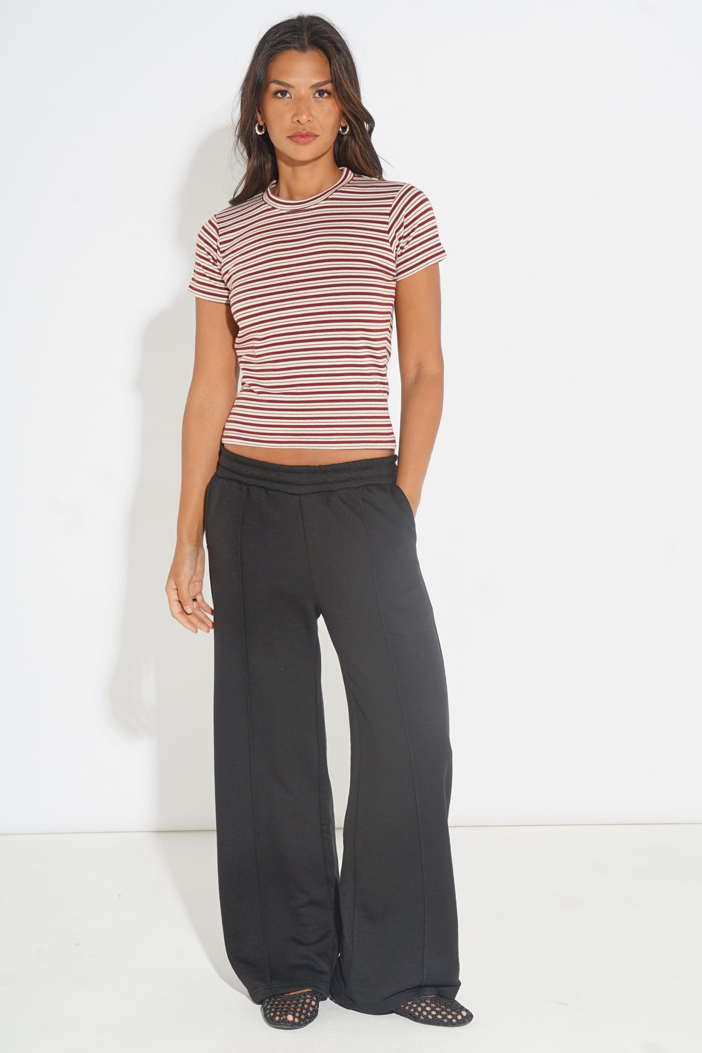 A woman modeling a short-sleeve cropped top with thin red and white horizontal stripes, paired with wide-leg black pants and black open-toe shoes, against a plain white studio background.
