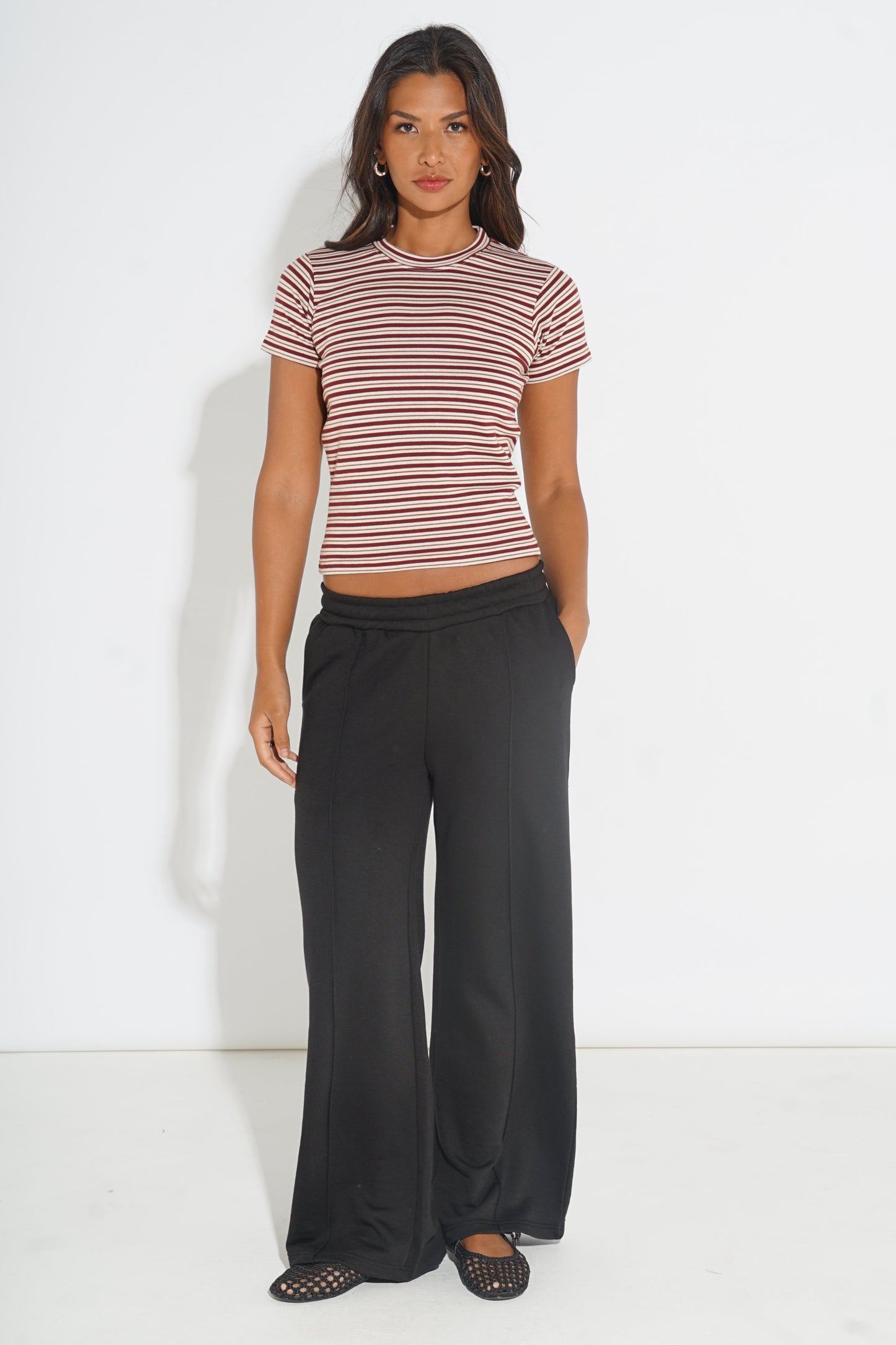 A woman modeling a short-sleeve striped crop top in red and white, paired with black wide-leg pants and black flat shoes, standing against a plain white studio background.