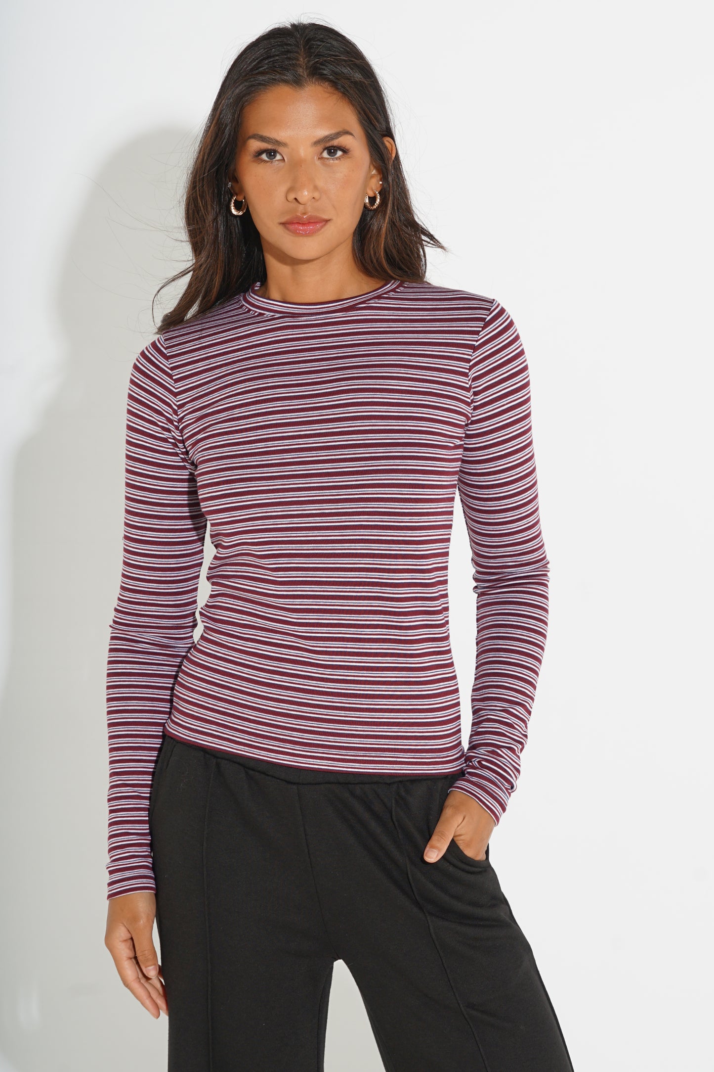 A woman modeling a long-sleeve crewneck top with thin horizontal stripes in dark red and purple, paired with black pants, against a plain white studio background.