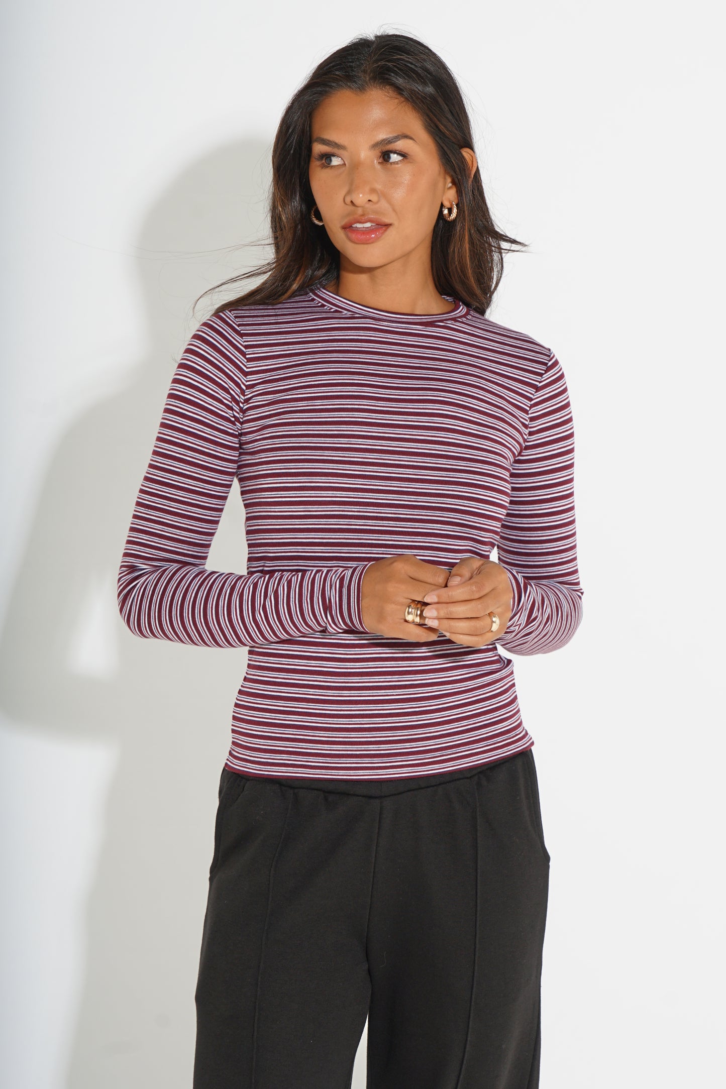 A woman modeling a long-sleeve crewneck top with thin horizontal stripes in dusty purple and burgundy, paired with black pants, against a plain white studio background.