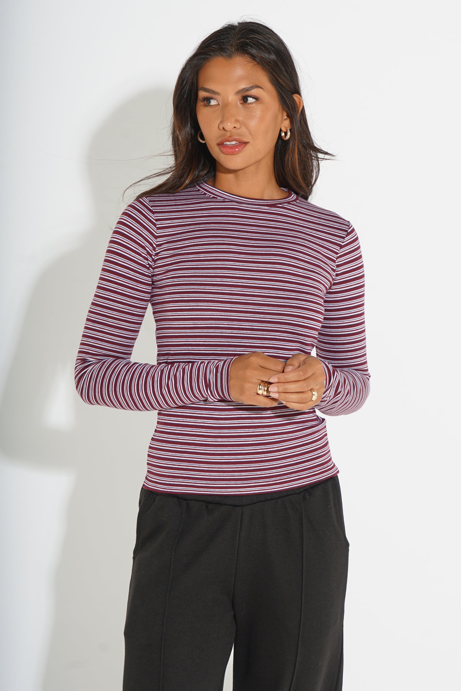 A woman modeling a long-sleeve crewneck top with thin horizontal stripes in dusty purple and burgundy, paired with black pants, against a plain white studio background.