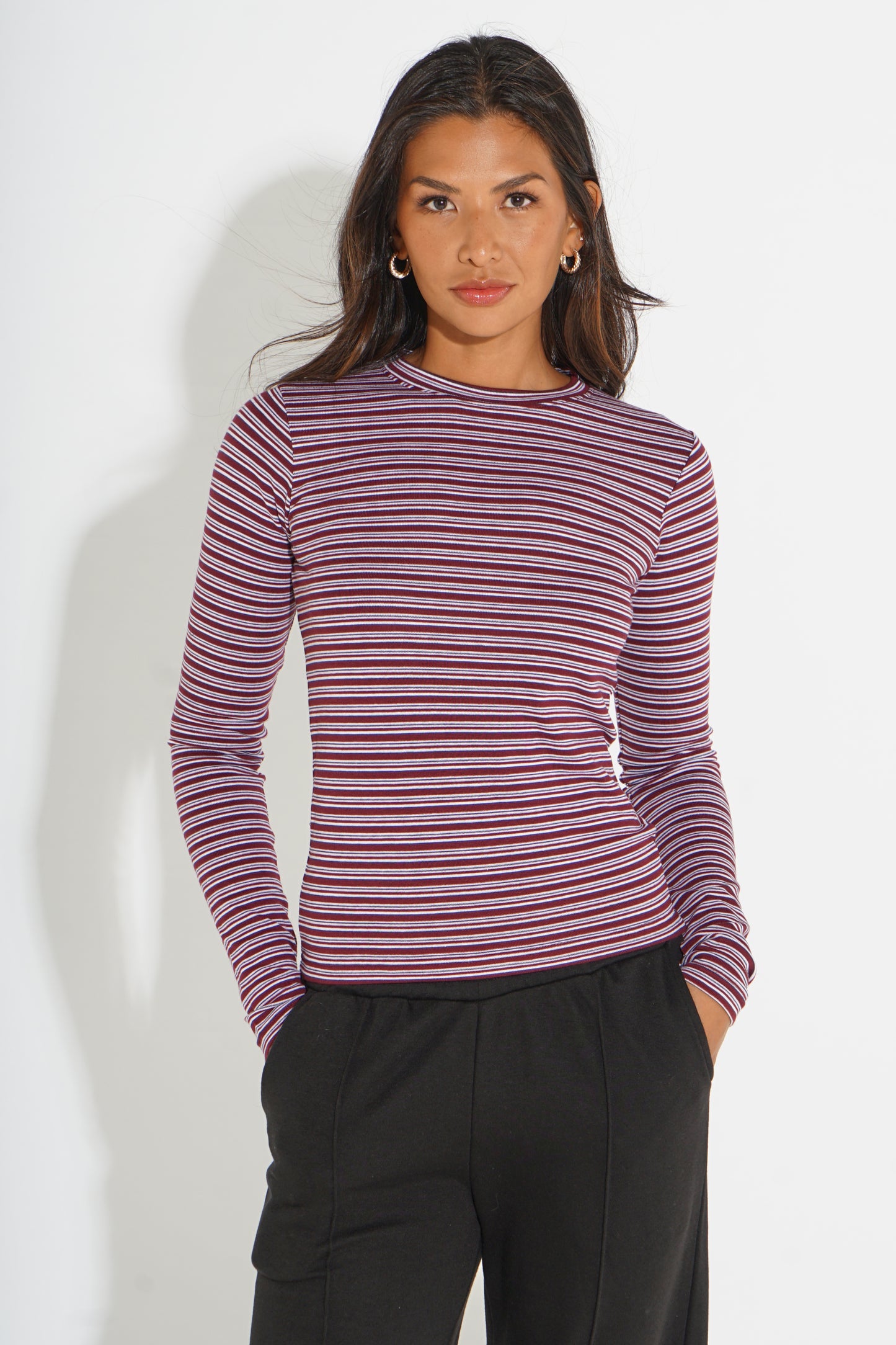 A woman modeling a long-sleeve crewneck top with horizontal burgundy and navy stripes, paired with black pants, against a plain white studio background.