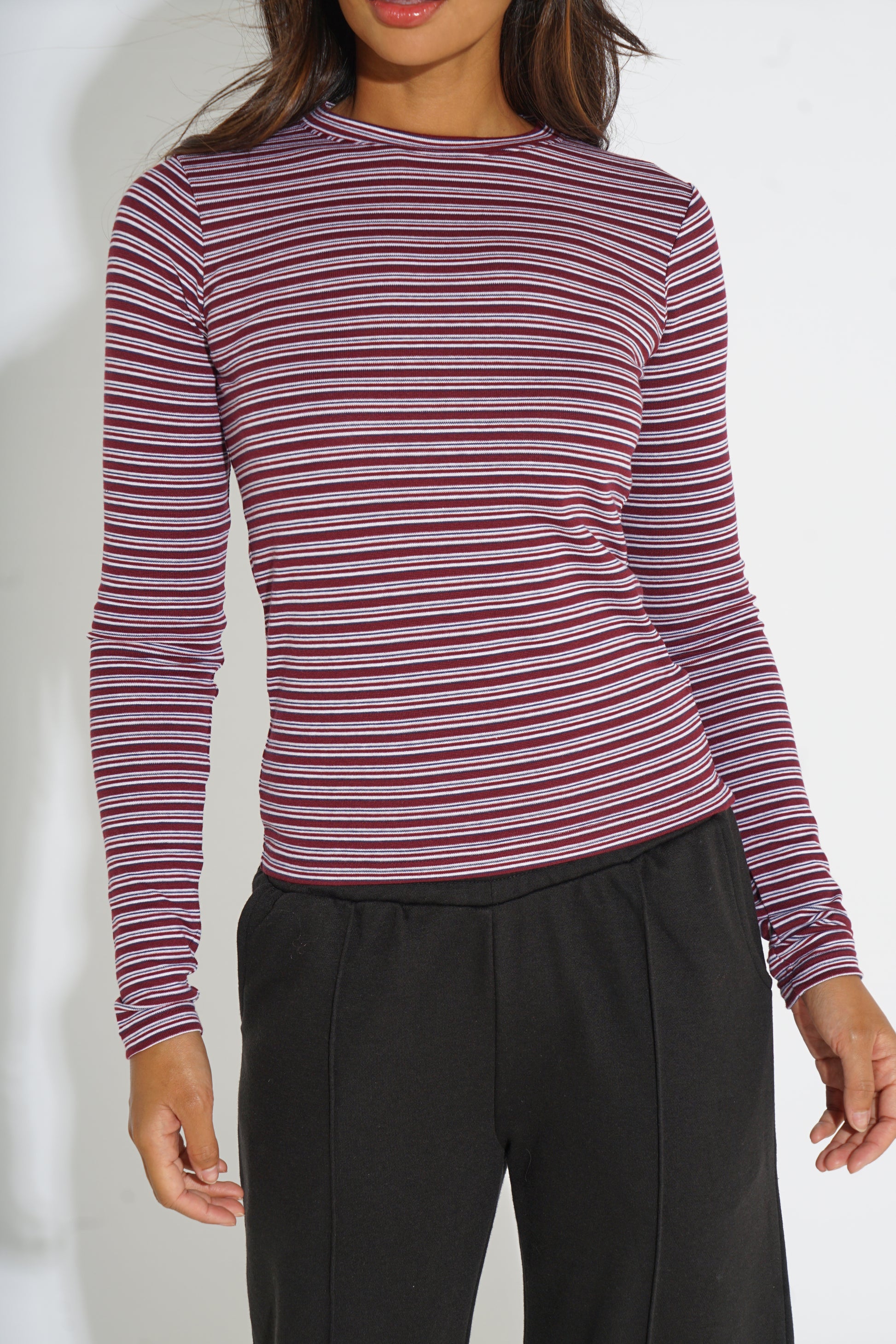 A woman wearing a long-sleeve crewneck top with thin horizontal stripes in burgundy, pink, and white, paired with black pants.