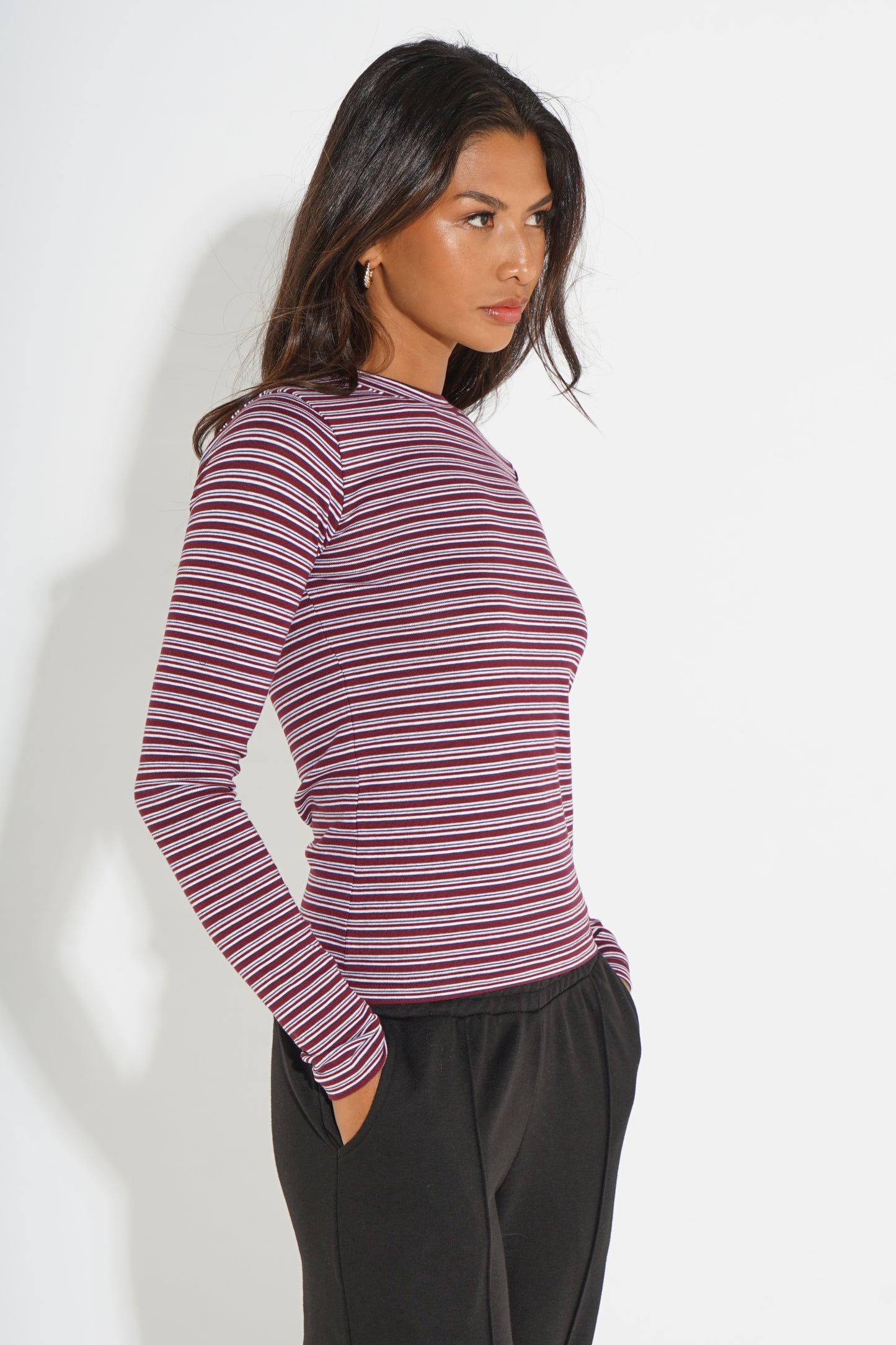 A woman wearing a long-sleeve ribbed knit top with horizontal burgundy and navy stripes, paired with black pants, photographed against a plain white studio background.