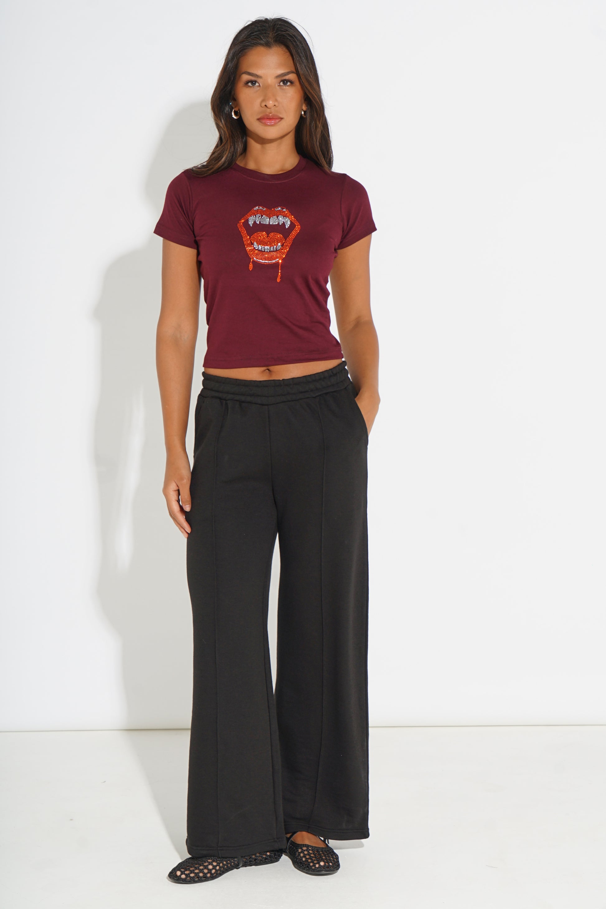 A woman modeling a maroon short-sleeve crop top with a colorful graphic print and black wide-leg pants, standing against a plain white studio background.