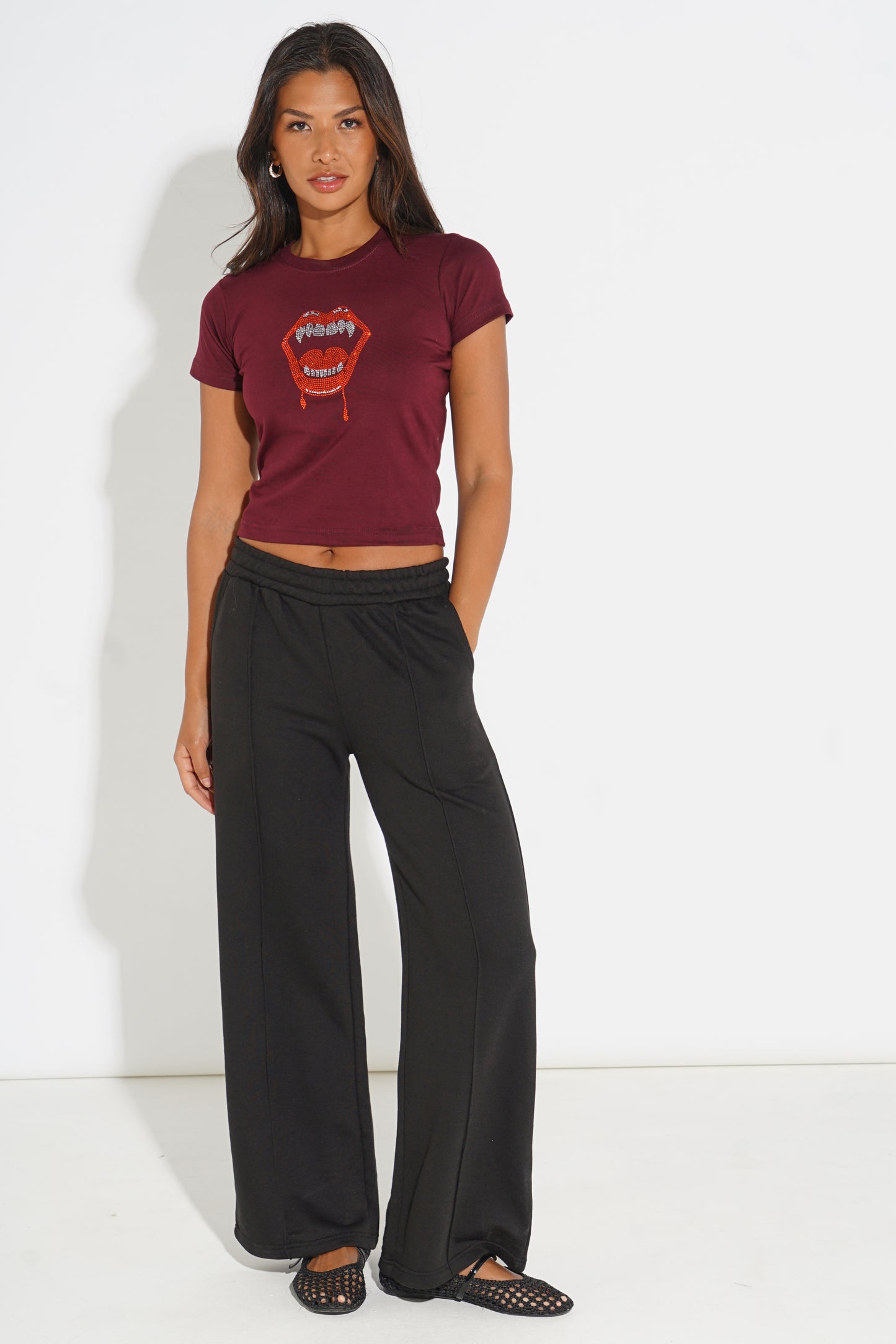 A woman modeling a maroon short-sleeve crop top with a red lips graphic and black wide-leg pants, standing against a plain white studio background.