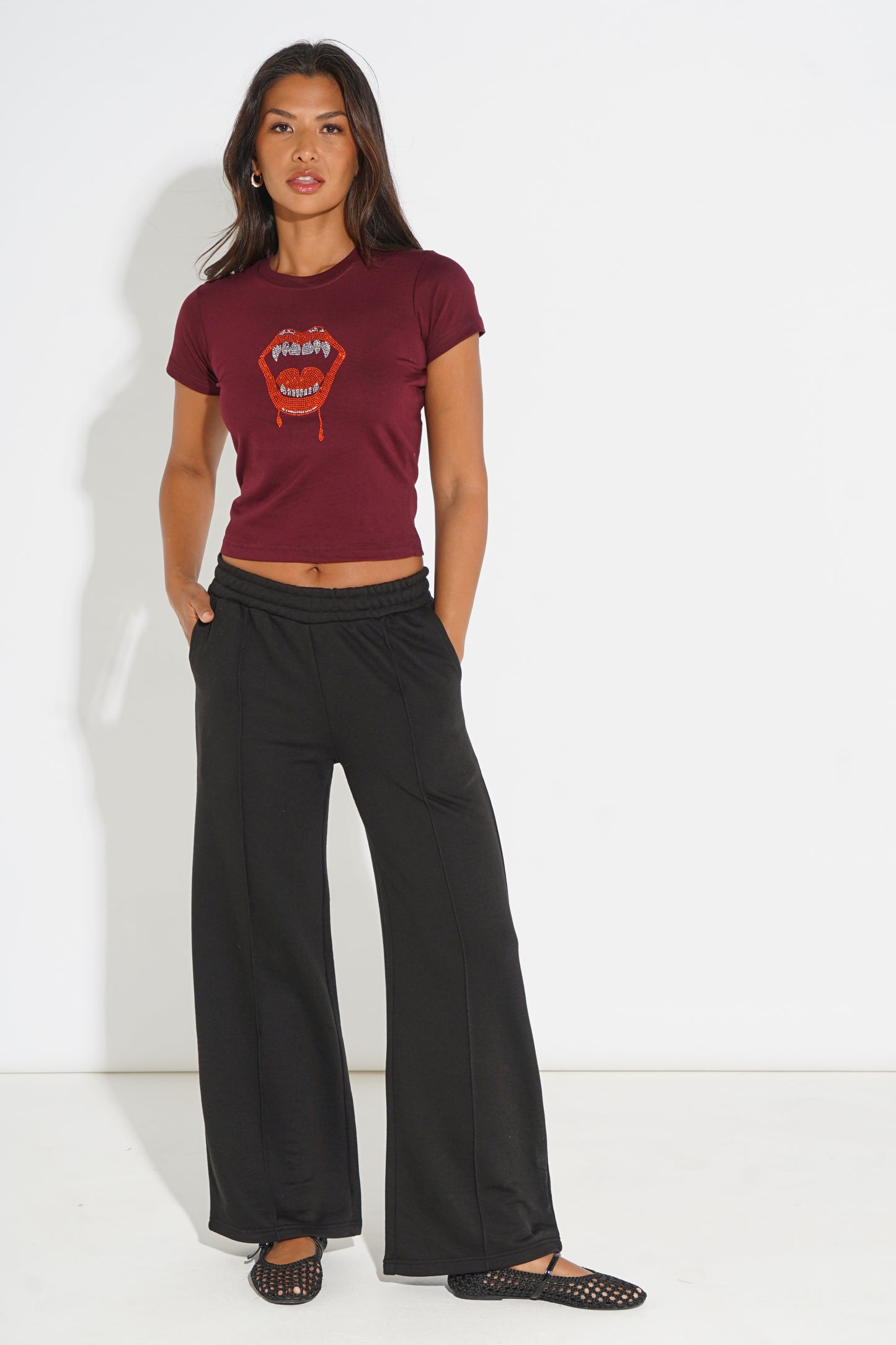 A woman modeling a short-sleeve maroon t-shirt with a red and orange graphic print on the front, paired with black wide-leg pants and black flat shoes, standing against a white studio background.