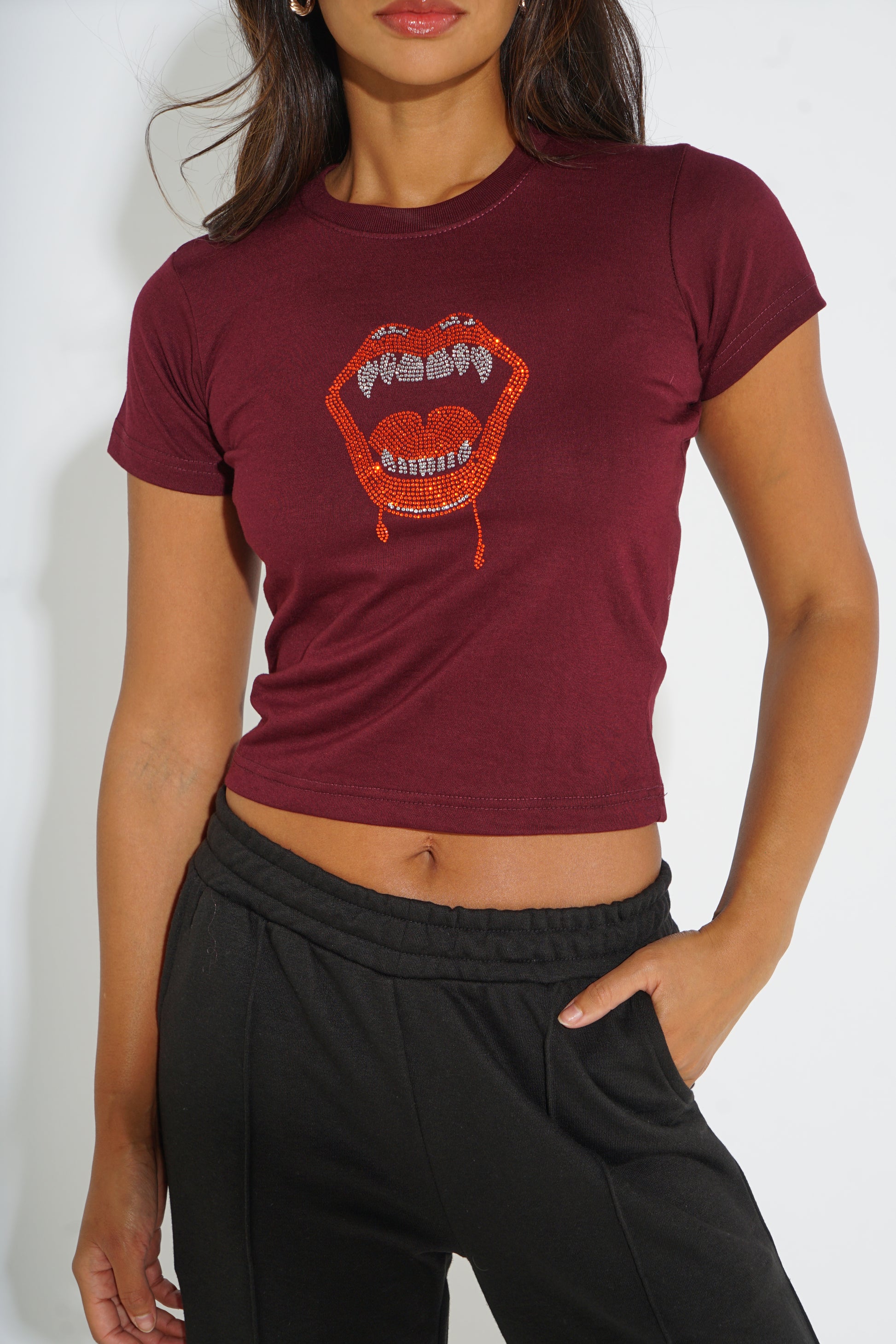 A woman wearing a maroon short-sleeve crop top with a red lips graphic on the front, paired with black sweatpants, against a plain white background.