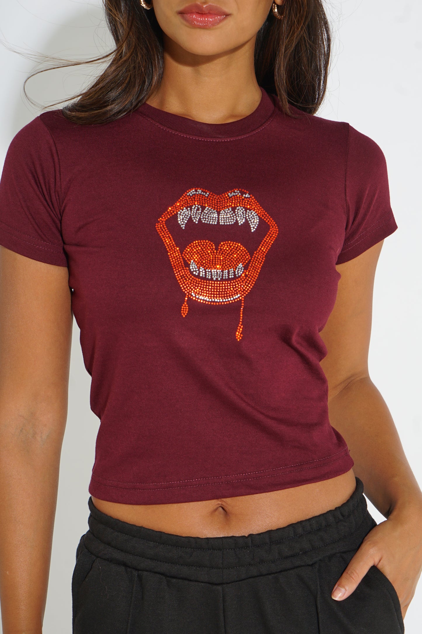 A woman wearing a maroon short-sleeve crop top with a rhinestone-embellished open mouth graphic on the front, paired with black sweatpants.