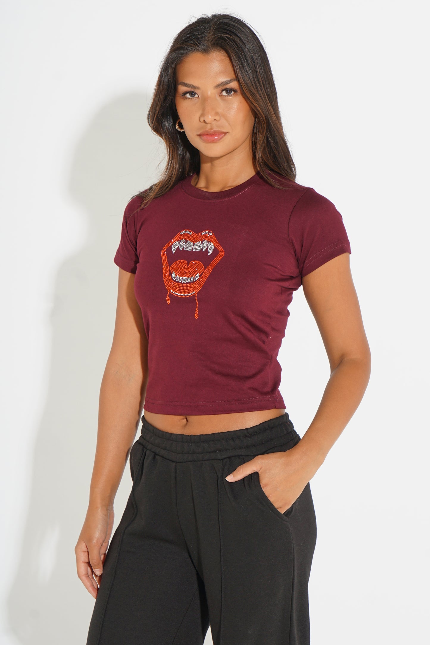 A woman modeling a short-sleeve maroon crop top with a graphic of dripping red lips and the word 'fassy' in white, paired with black sweatpants against a white studio background.