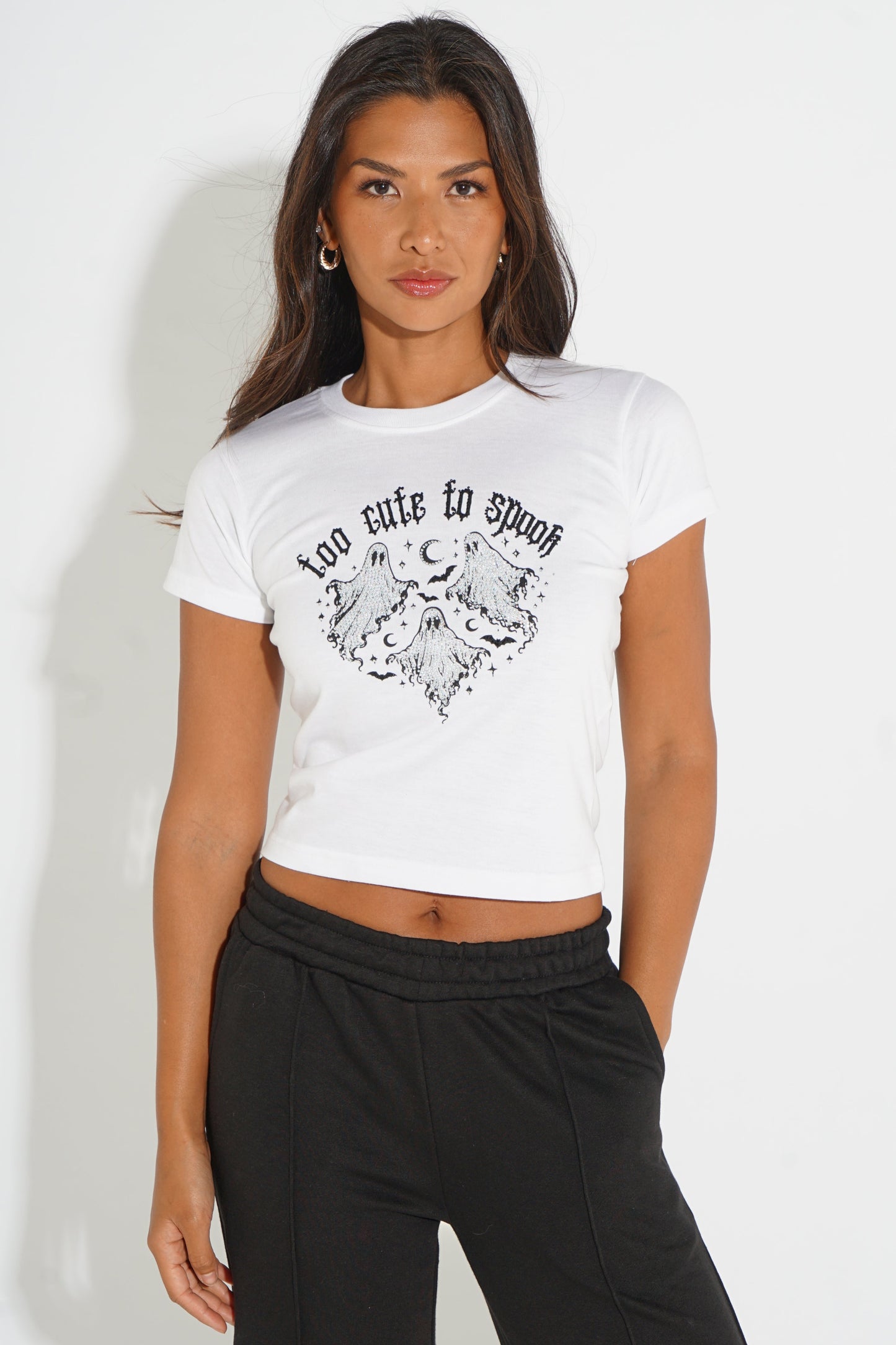 A woman modeling a white cropped short-sleeve t-shirt with a black graphic print of a winged heart and the text 'too cute to speak' on the front, paired with black sweatpants against a plain white background.