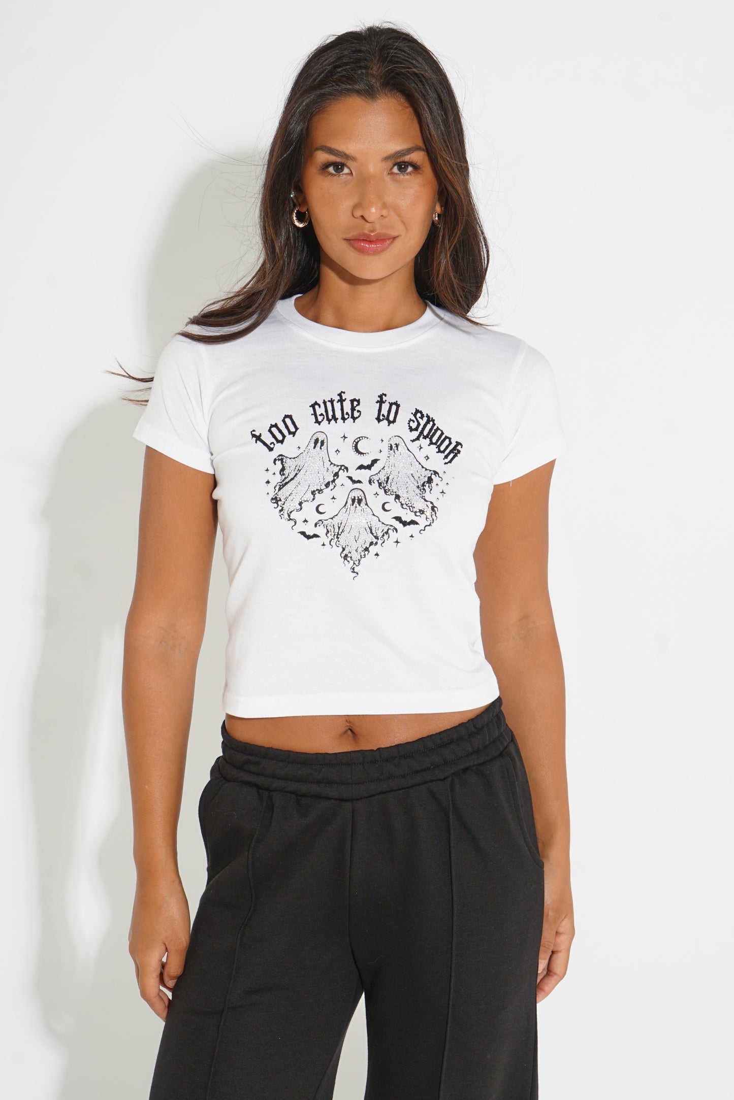A woman modeling a white short-sleeve crop top with a black graphic print that reads 'too cute to spoil' above a moon and cloud illustration, paired with black sweatpants against a plain white background.