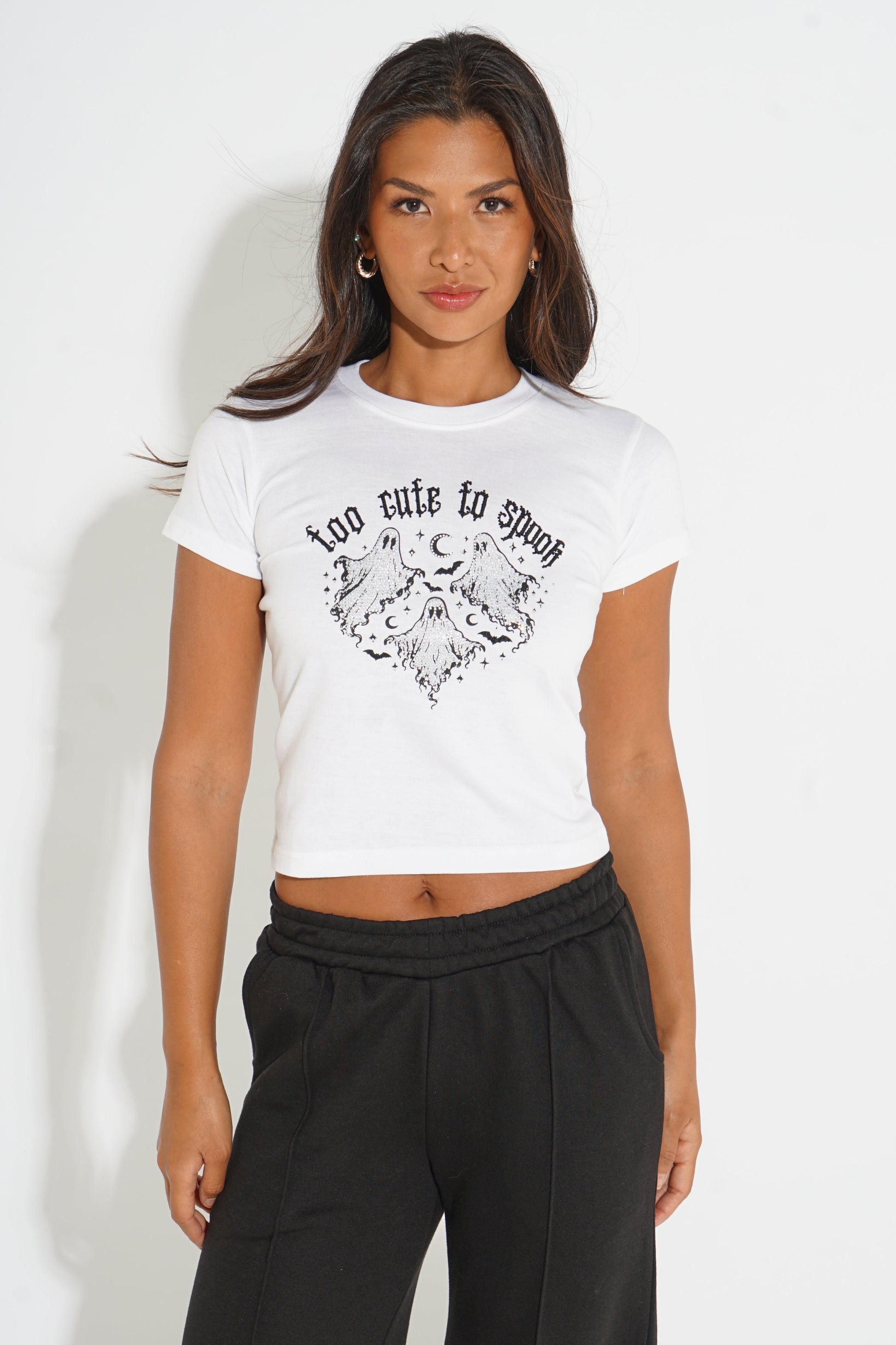 A woman modeling a white short-sleeve crop top with a black graphic print that reads 'too cute to spoil' above a moon and cloud illustration, paired with black sweatpants against a plain white background.