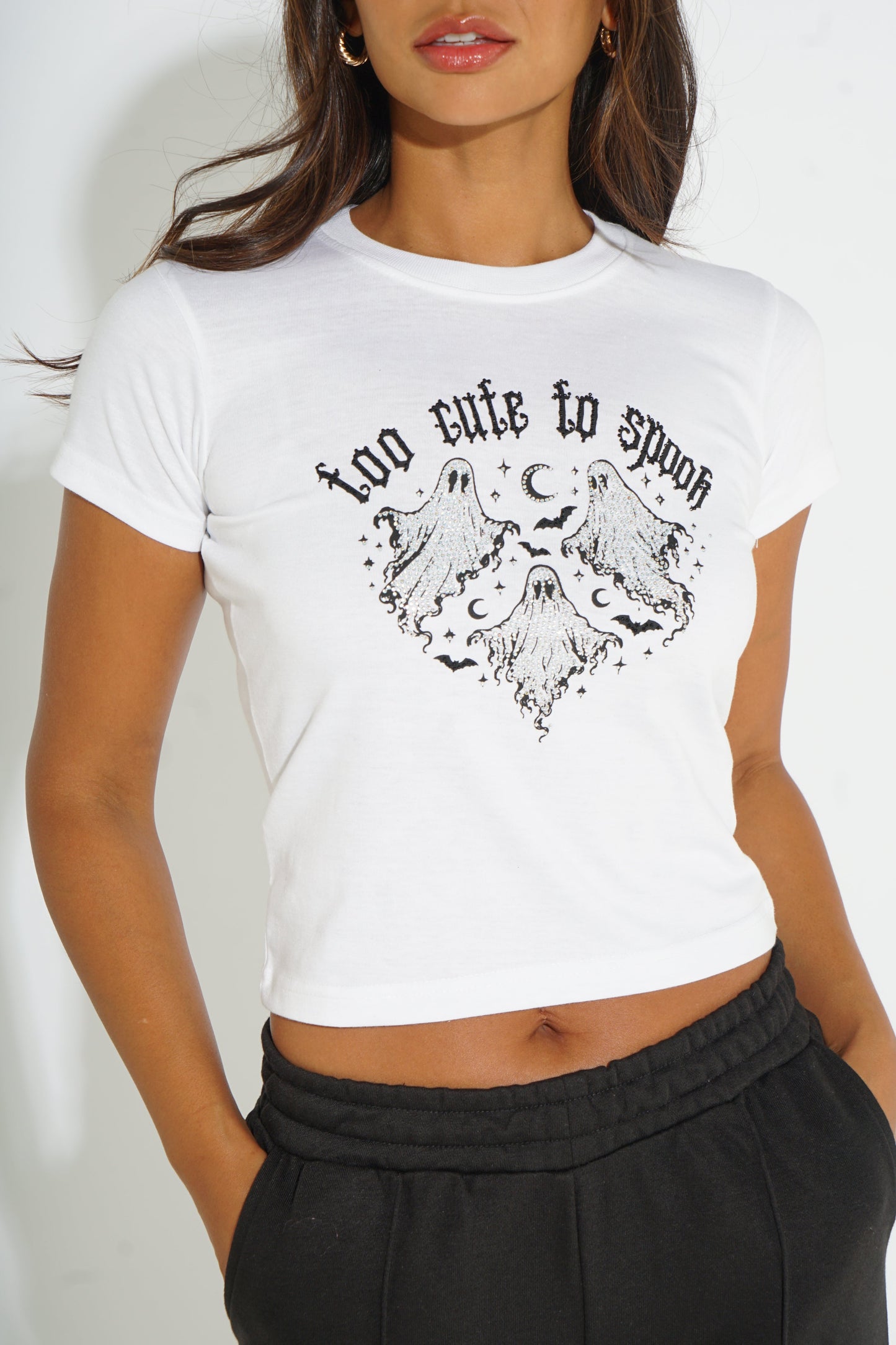 A woman wearing a white cropped t-shirt with a black graphic print that reads 'too cute to spoon' above a heart-shaped ghost illustration, paired with black sweatpants.