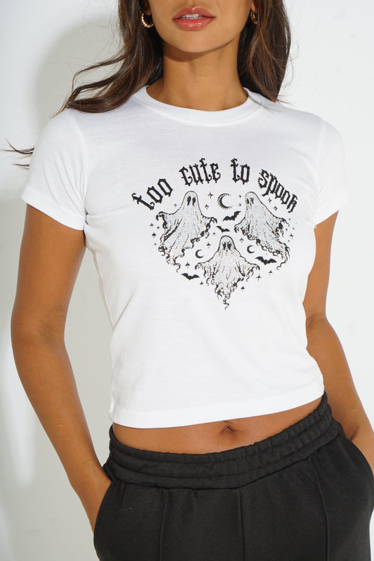 A woman wearing a white cropped t-shirt with a black graphic print that reads 'too cute to spoon' above a heart-shaped ghost illustration, paired with black sweatpants.