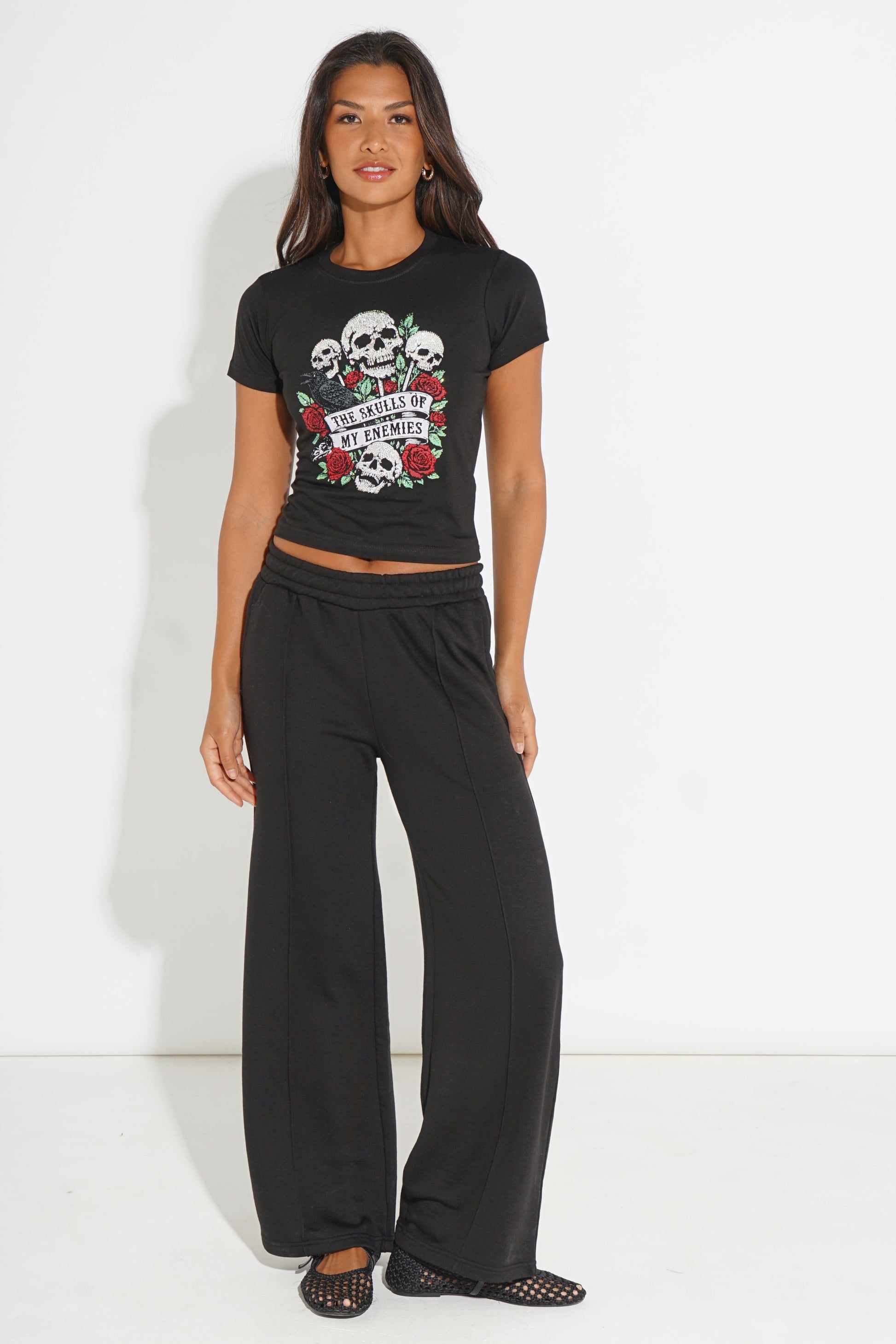 A woman modeling a black short-sleeve crop top with a skull and rose graphic, paired with black wide-leg pants and black open-toe mule sandals, against a plain white studio background.