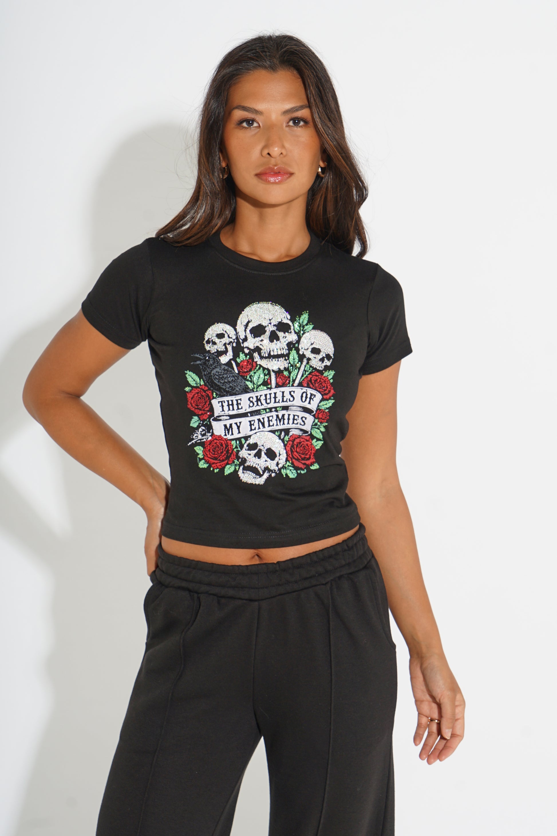 A woman modeling a black short-sleeve crop top with a graphic print of skulls, red roses, and a banner reading 'THE SKULLS OF MY ENEMIES'. She is also wearing black sweatpants against a plain white studio background.