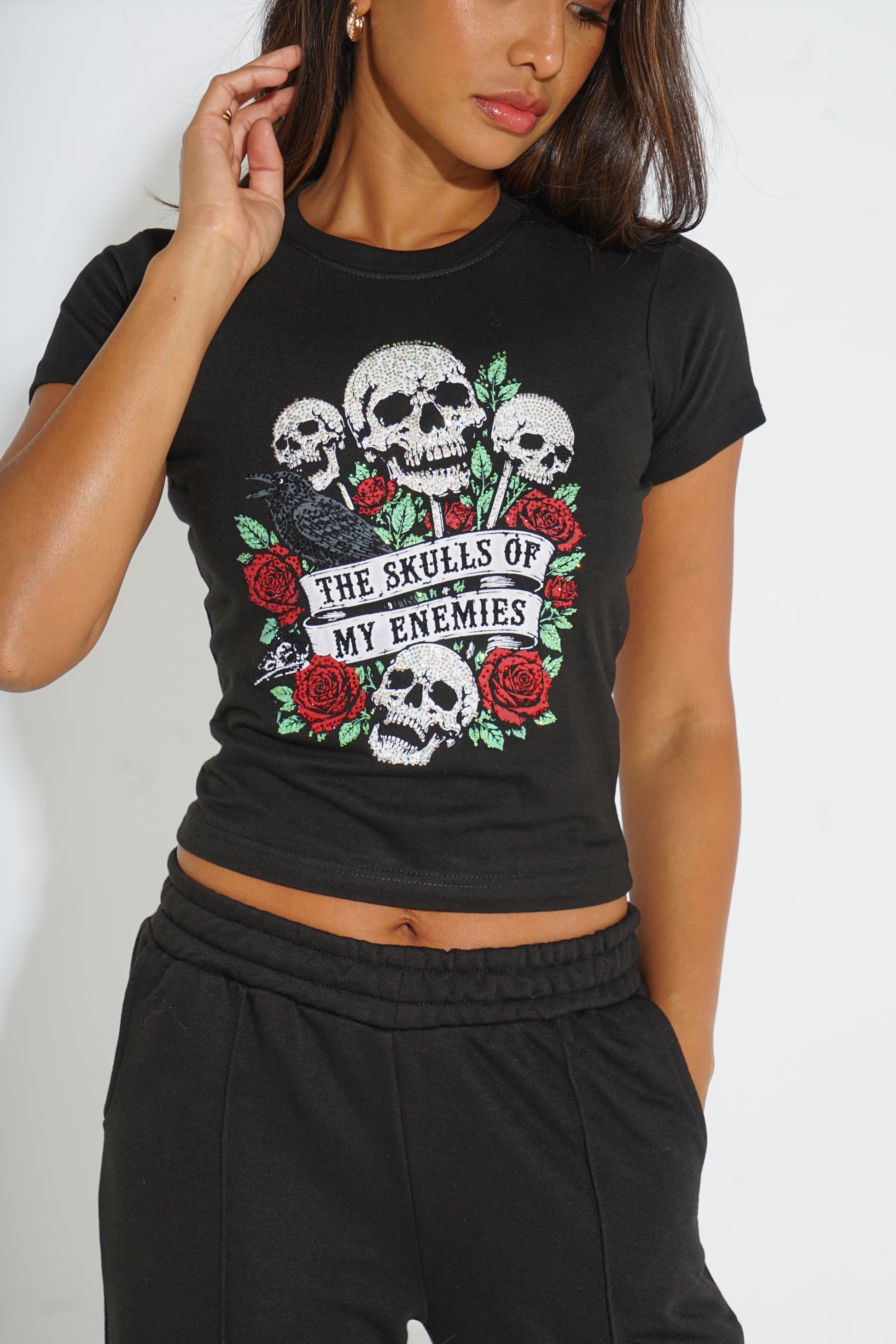 A woman wearing a black short-sleeve crop top t-shirt with a graphic print of skulls, red roses, and a black bird with the text 'THE SKULLS OF MY ENEMIES'.