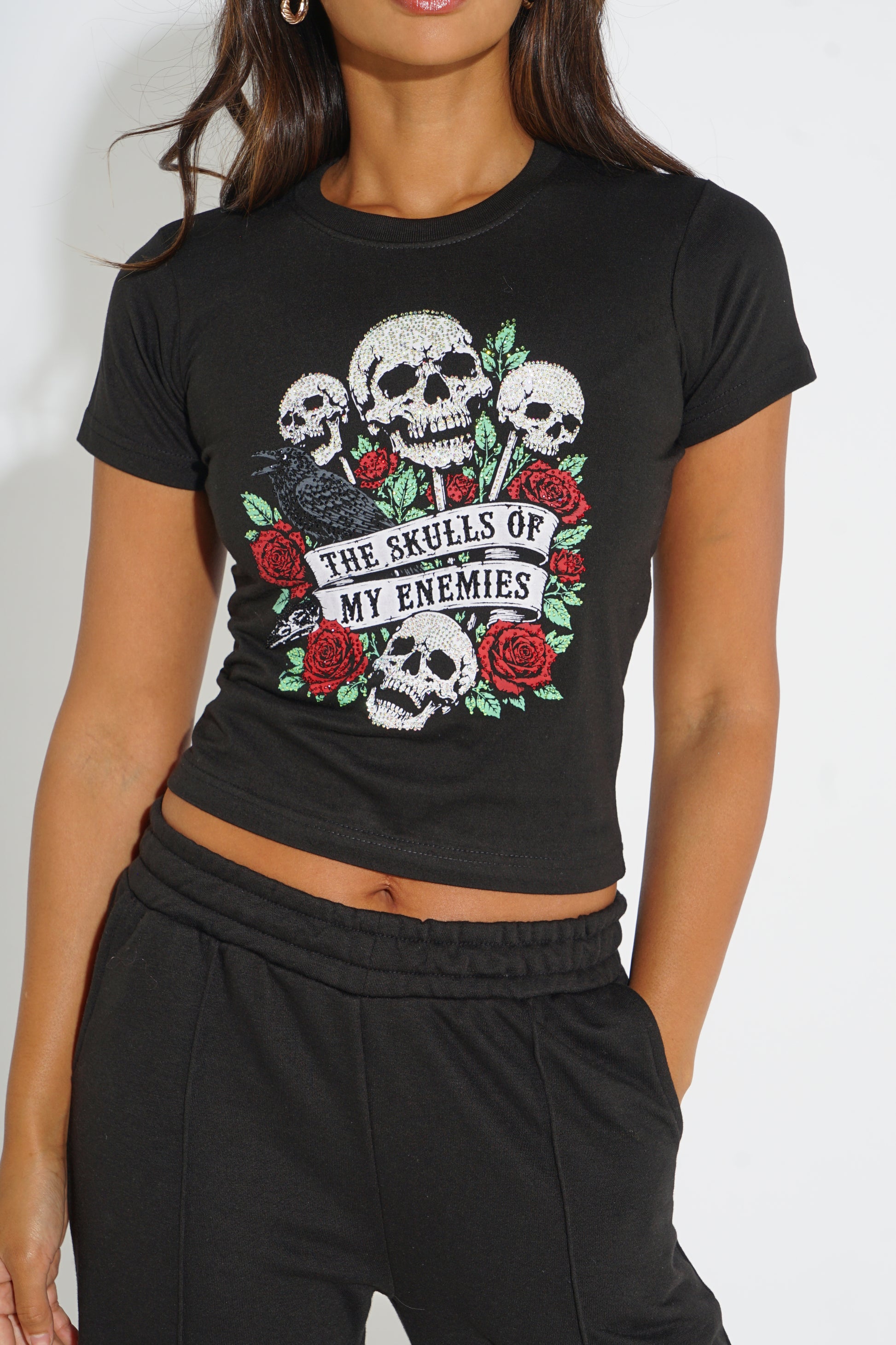 A woman wearing a black cropped t-shirt with a graphic of skulls, red roses, and a blackbird with the text 'THE SKULLS OF MY ENEMIES'.