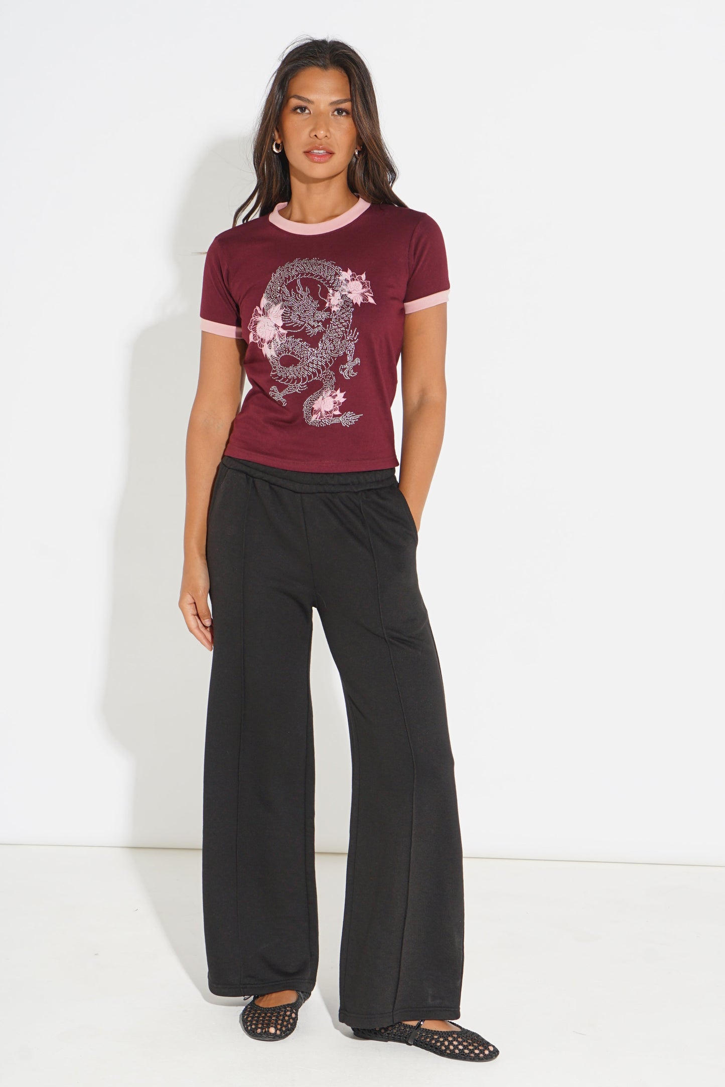 A woman modeling a short-sleeve maroon t-shirt with a pink and white dragon graphic, paired with black wide-leg pants and black flat shoes, standing against a white studio background.