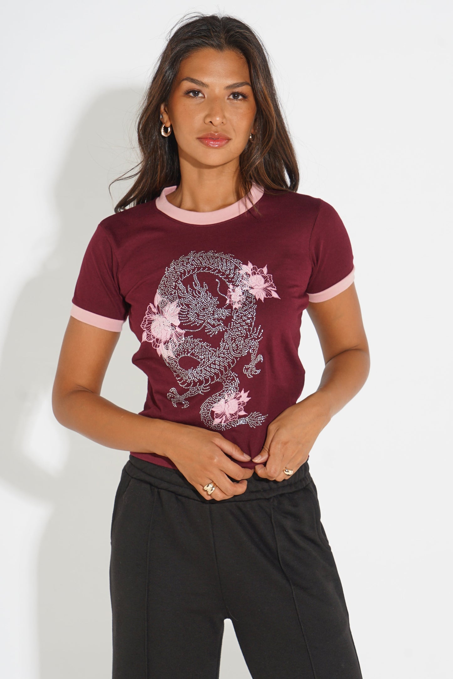A woman modeling a short-sleeve ringer t-shirt in burgundy with light pink trim and a large silver dragon graphic with pink flowers on the front, paired with black sweatpants against a white studio background.