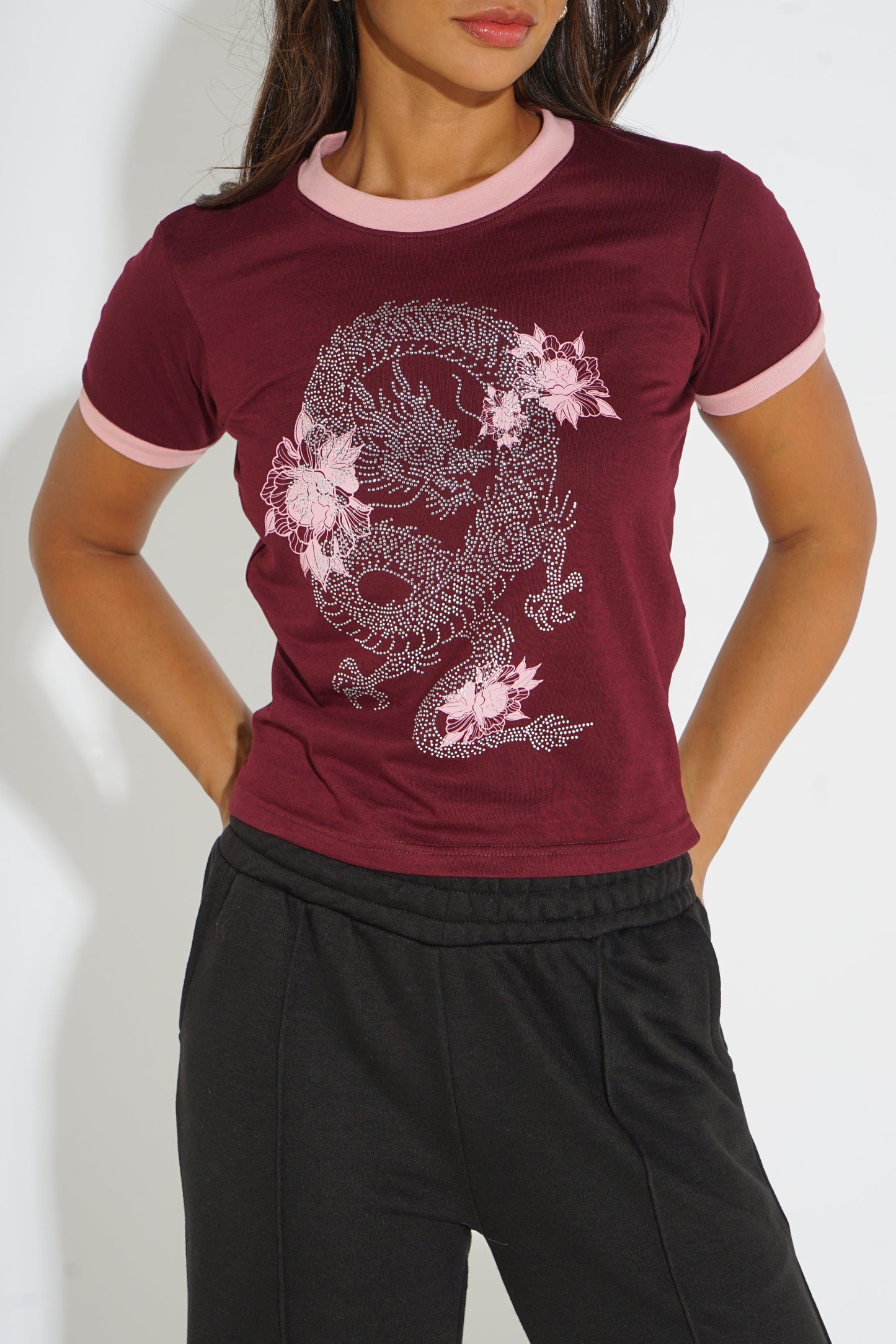 A woman wearing a short-sleeve ringer t-shirt in maroon with light pink trim at the collar and sleeve cuffs, featuring a large white dragon graphic with pink flowers on the front. She is also wearing black sweatpants against a plain white background.