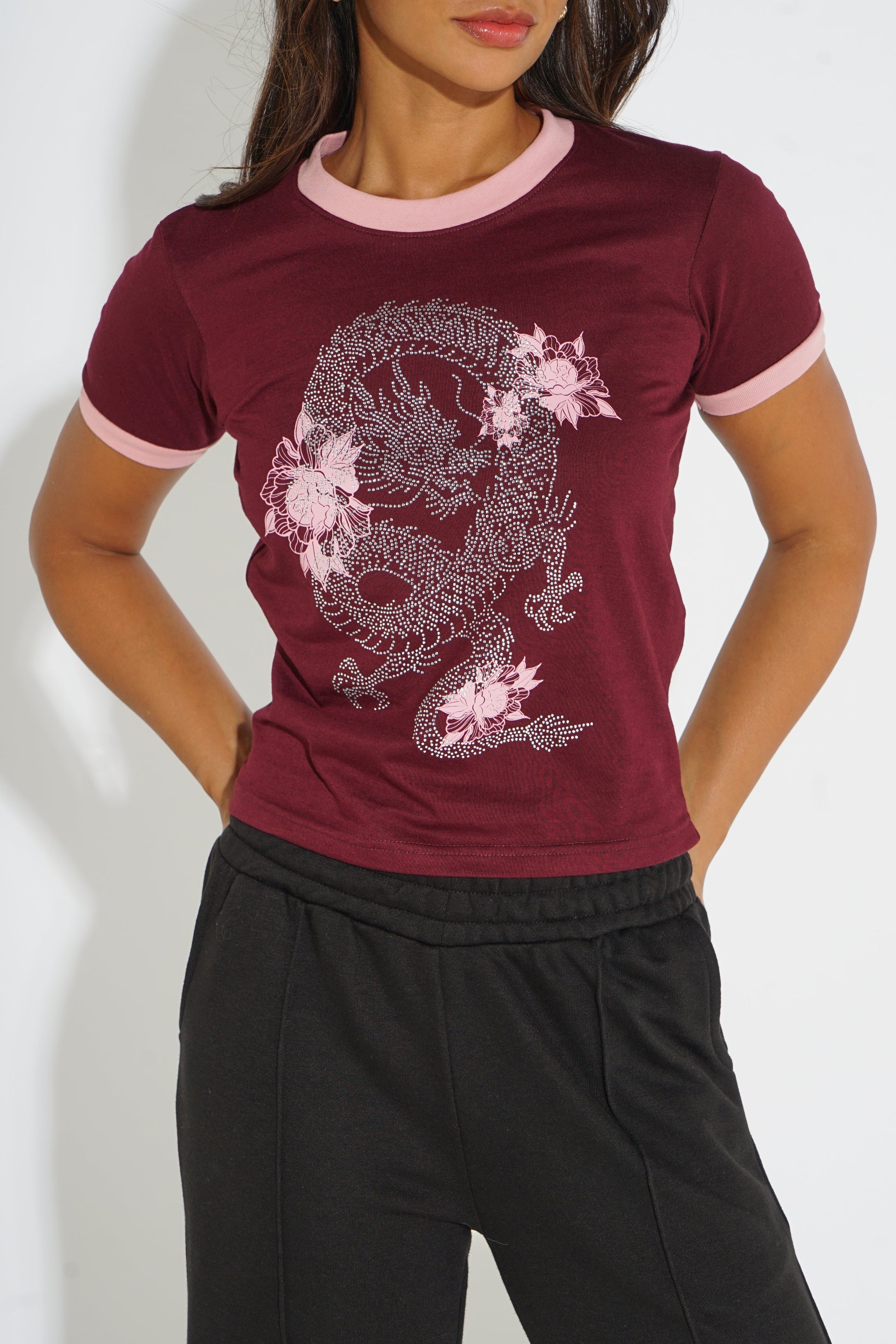 A woman wearing a short-sleeve ringer t-shirt in maroon with light pink trim at the collar and sleeve cuffs, featuring a large white dragon graphic with pink flowers on the front. She is also wearing black sweatpants against a plain white background.