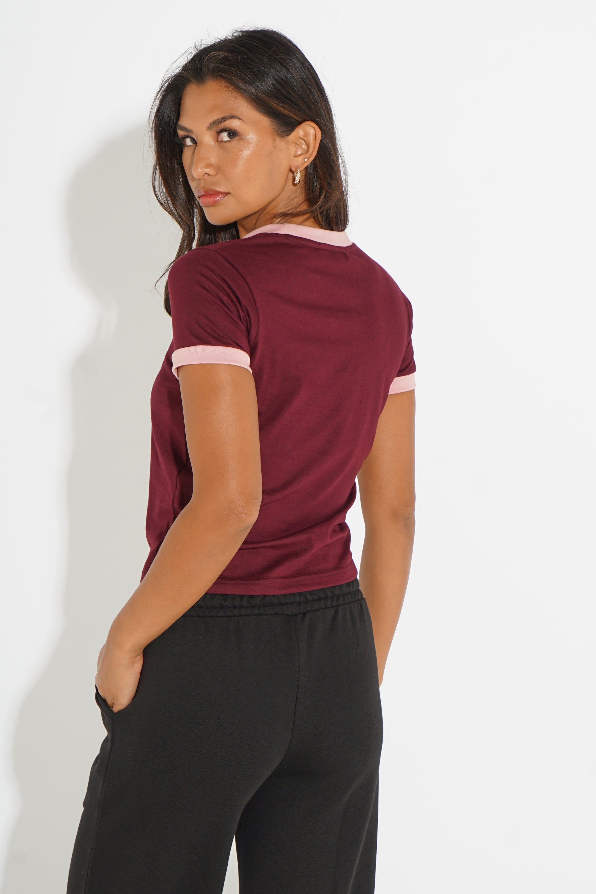 A woman wearing a short-sleeve ringer t-shirt in burgundy with light pink trim on the collar and sleeve cuffs, paired with black pants, photographed against a plain white wall.
