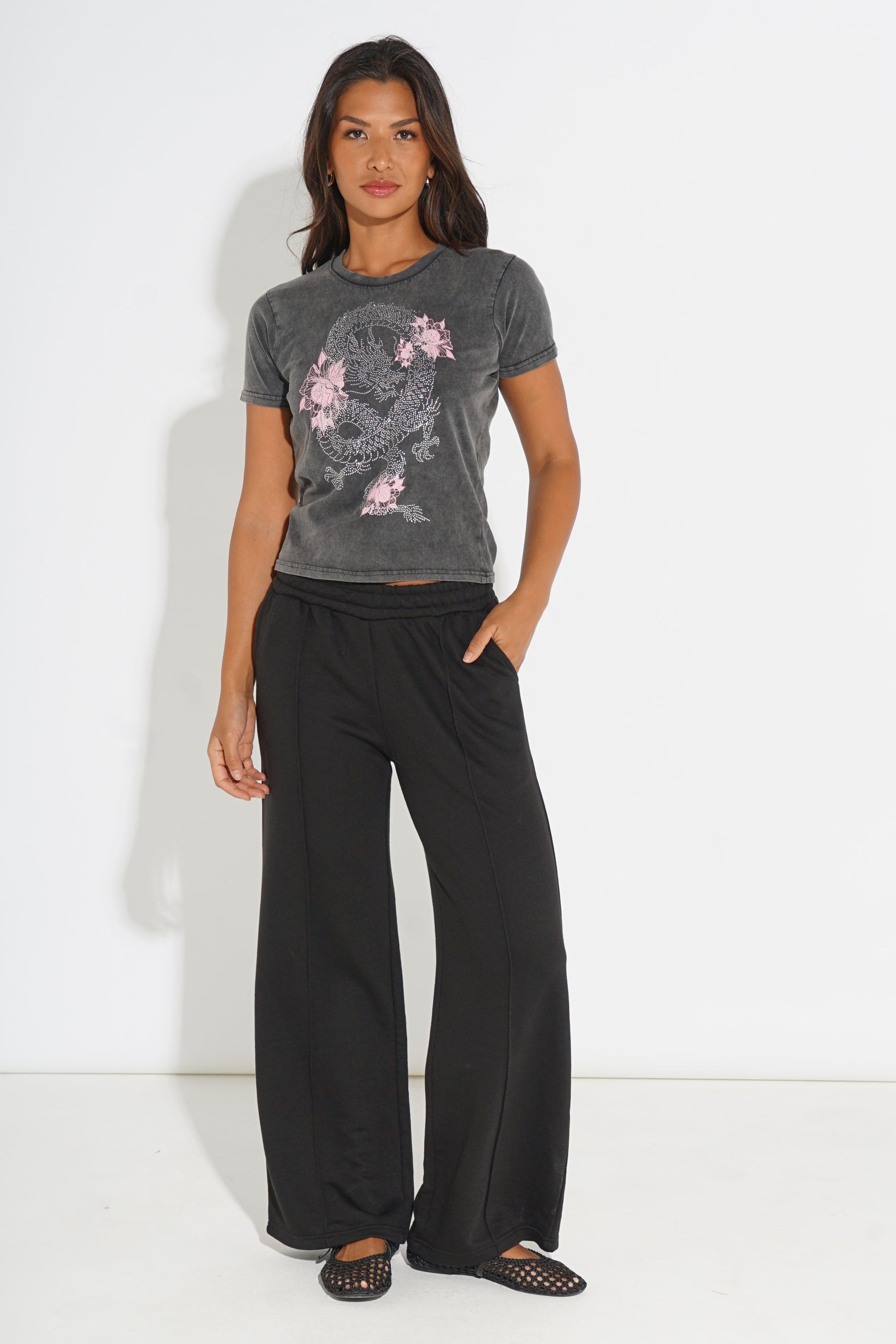 A woman modeling a dark gray washed graphic t-shirt with a pink dragon design, paired with black wide-leg pants and black flat sandals, standing against a white studio background.