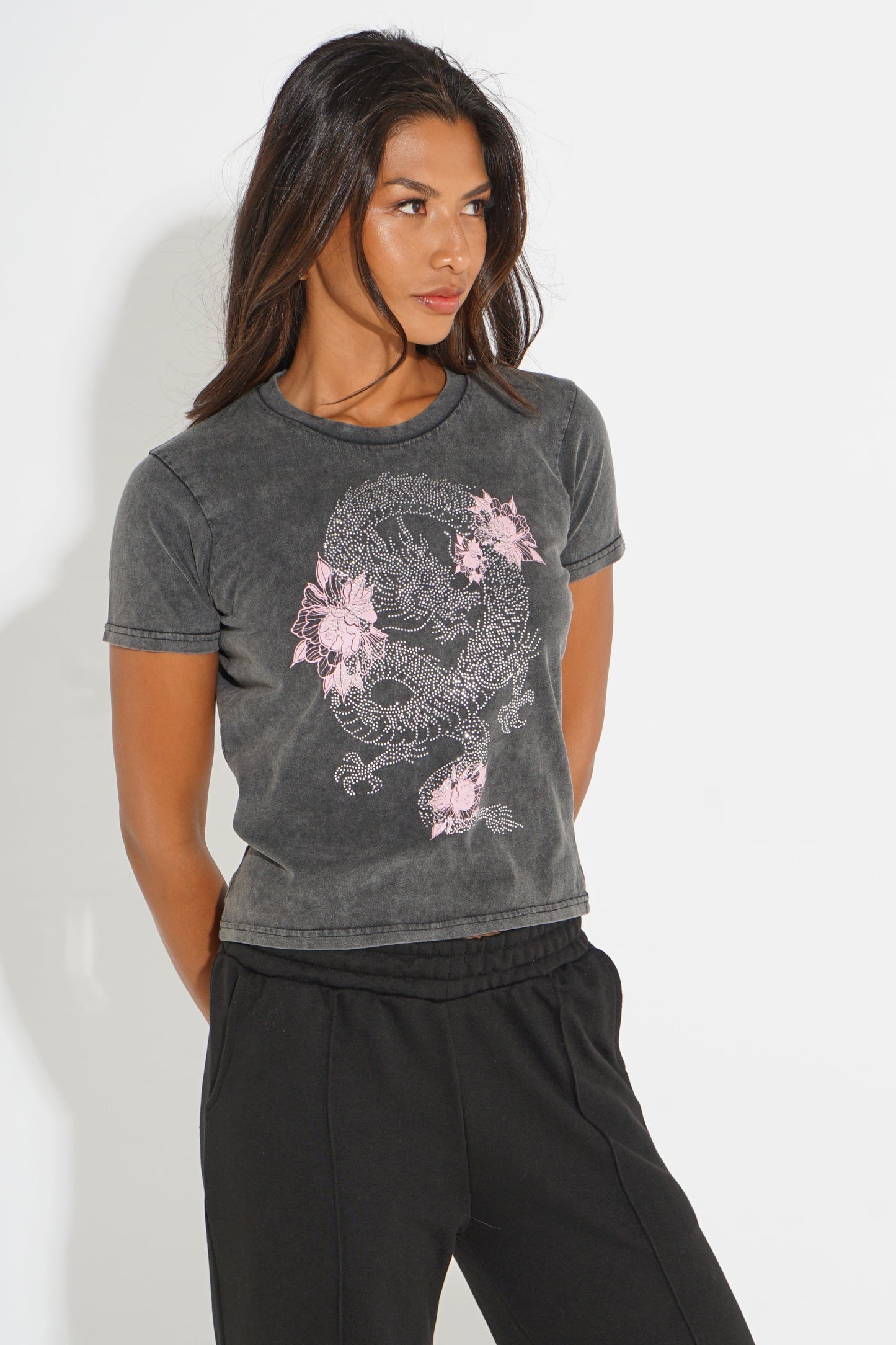 A woman modeling a dark gray washed short-sleeve t-shirt with a pink and white dragon graphic on the front, paired with black sweatpants against a white studio background.