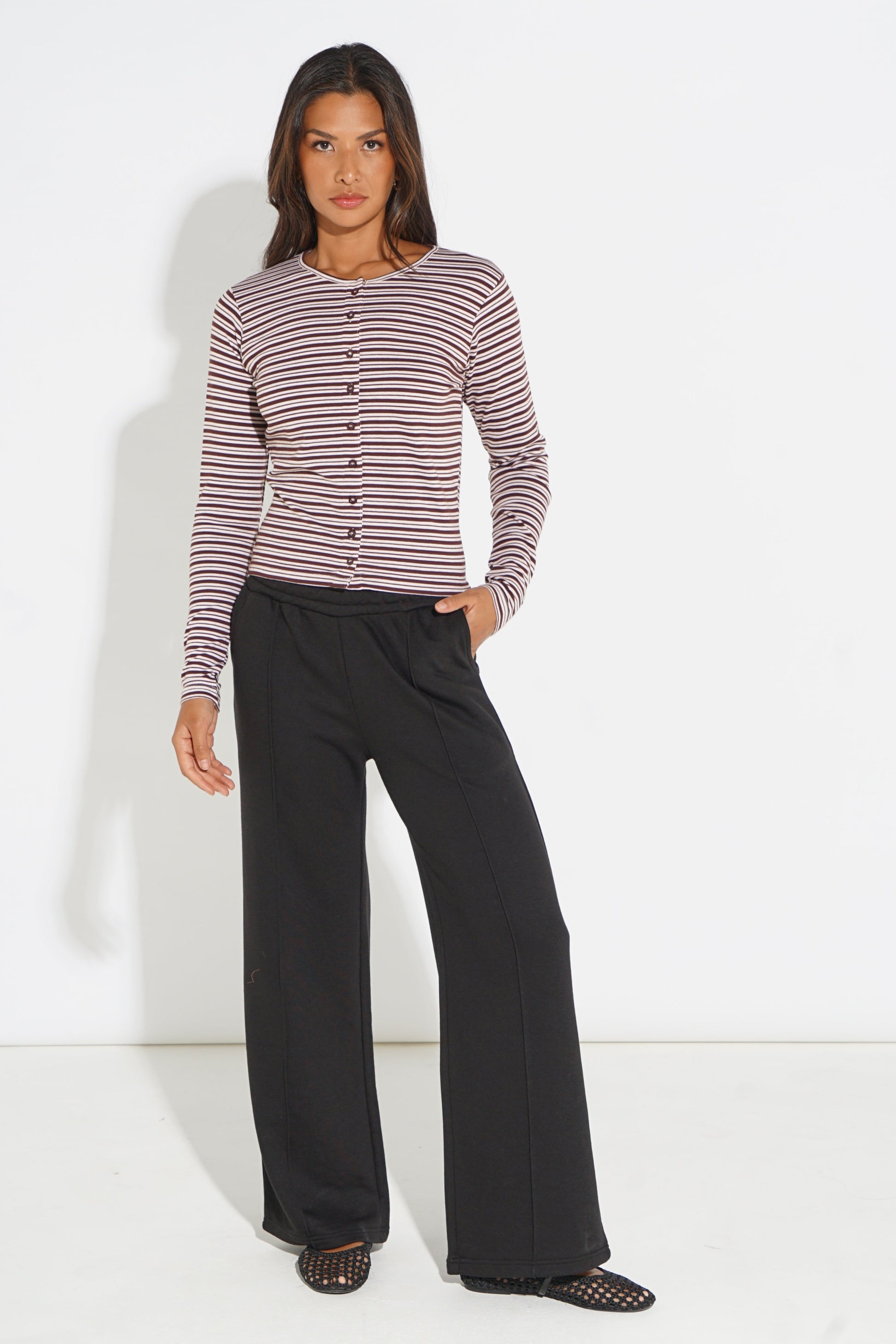 A woman modeling a long-sleeve button-front top with thin horizontal stripes in dusty pink and dark brown, paired with wide-leg black pants and black mesh flat shoes, against a plain white studio background.