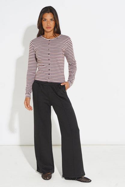 A woman modeling a long-sleeve button-front top with thin horizontal stripes in dusty pink and dark brown, paired with wide-leg black pants and black mesh flat shoes, against a plain white studio background.