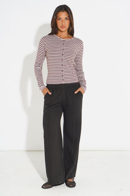 A woman modeling a long-sleeve knit top with thin horizontal stripes in dark red and off-white, paired with black wide-leg trousers and black flat shoes, against a plain white studio background.