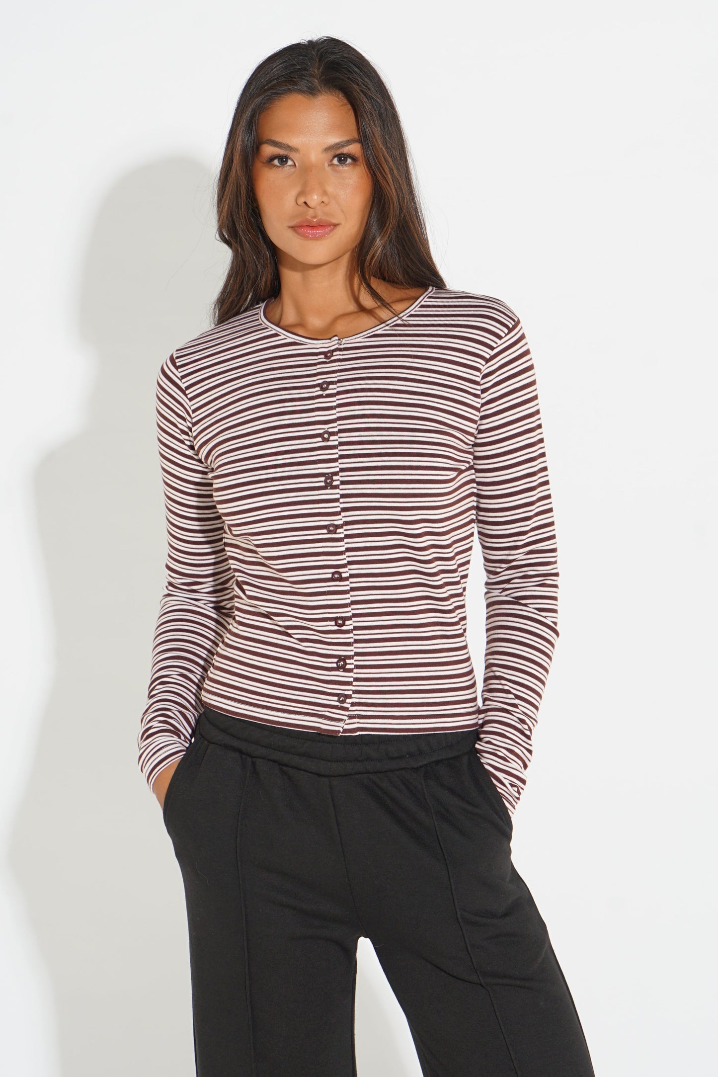 A woman modeling a long-sleeve button-front top with thin horizontal stripes in dark brown and muted pink, paired with black pants, against a plain light gray studio background.