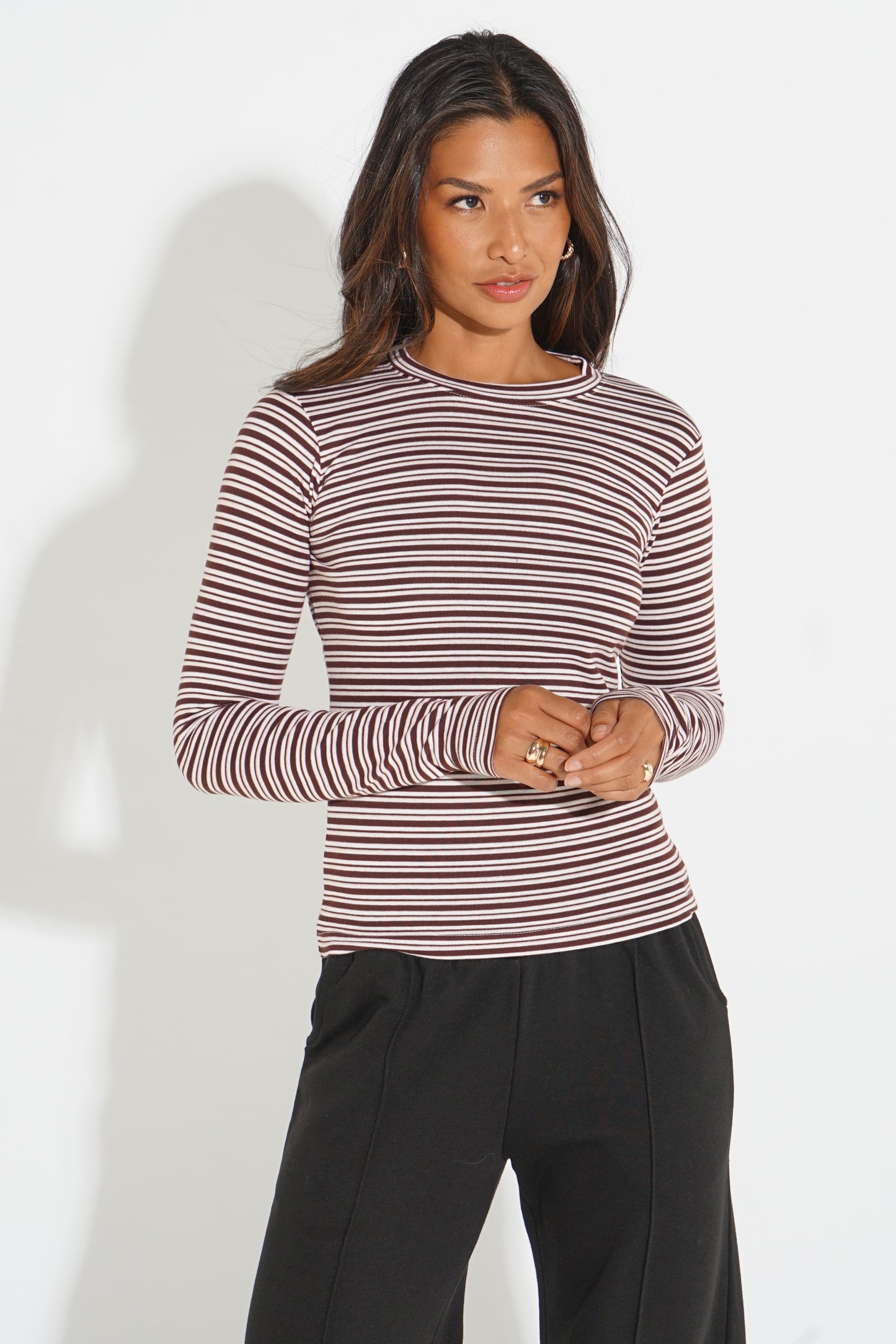 A woman modeling a long-sleeve crewneck top with thin horizontal stripes in dark red and beige, paired with black pants, against a plain white background.