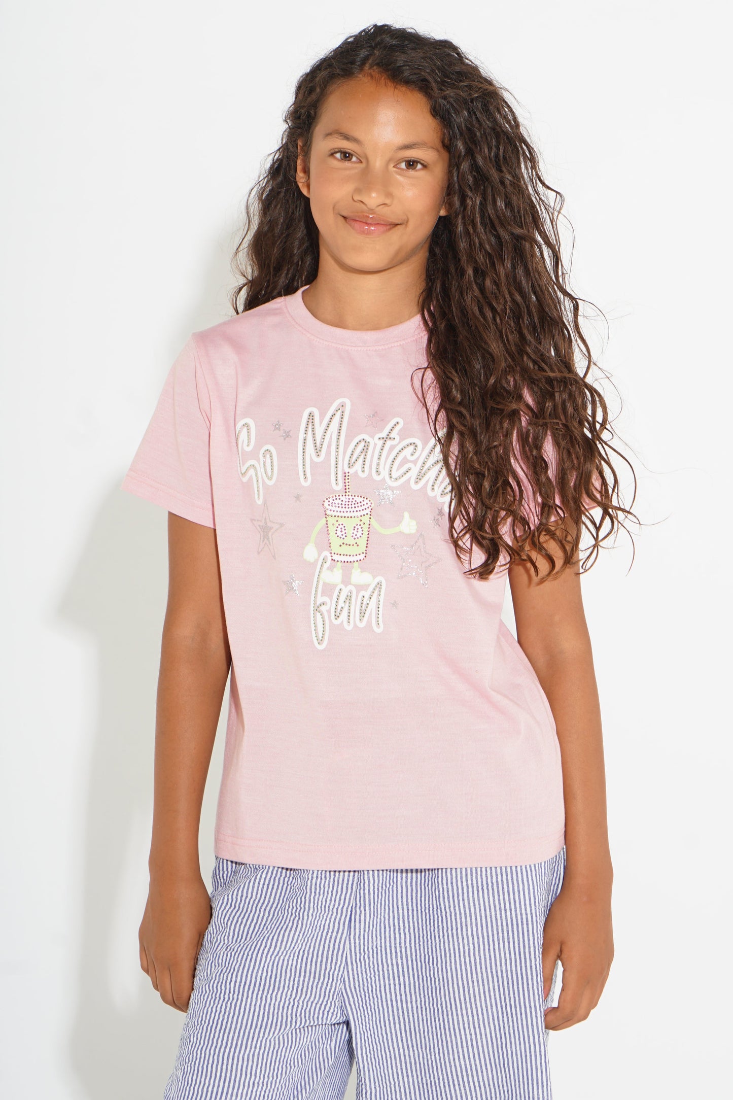 A young girl with long, curly brown hair wearing a light pink graphic t-shirt and blue-and-white striped pants, standing against a plain white background.