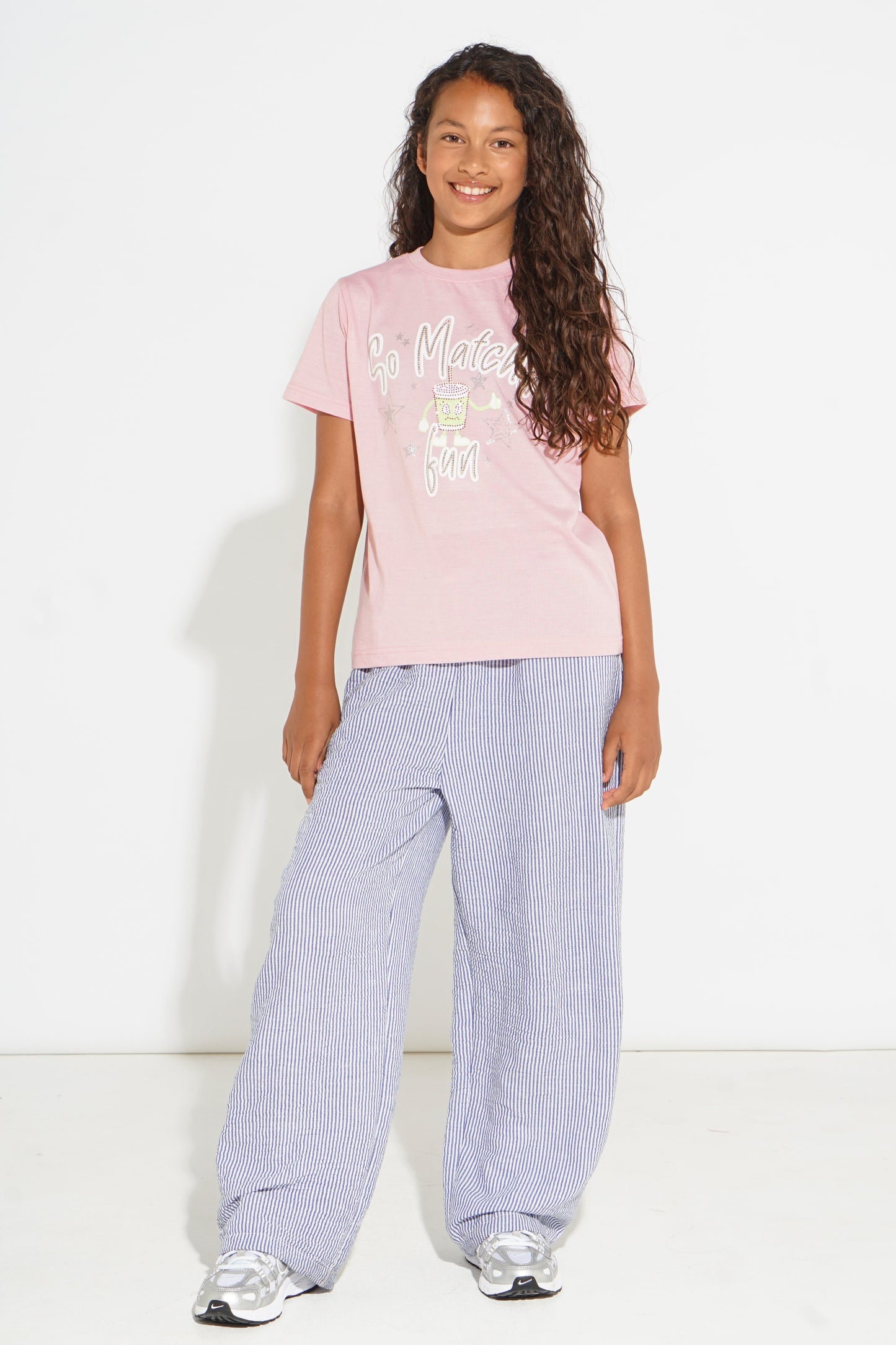 A young girl with long curly brown hair stands against a white studio background wearing a light pink graphic t-shirt and light blue wide-leg trousers.