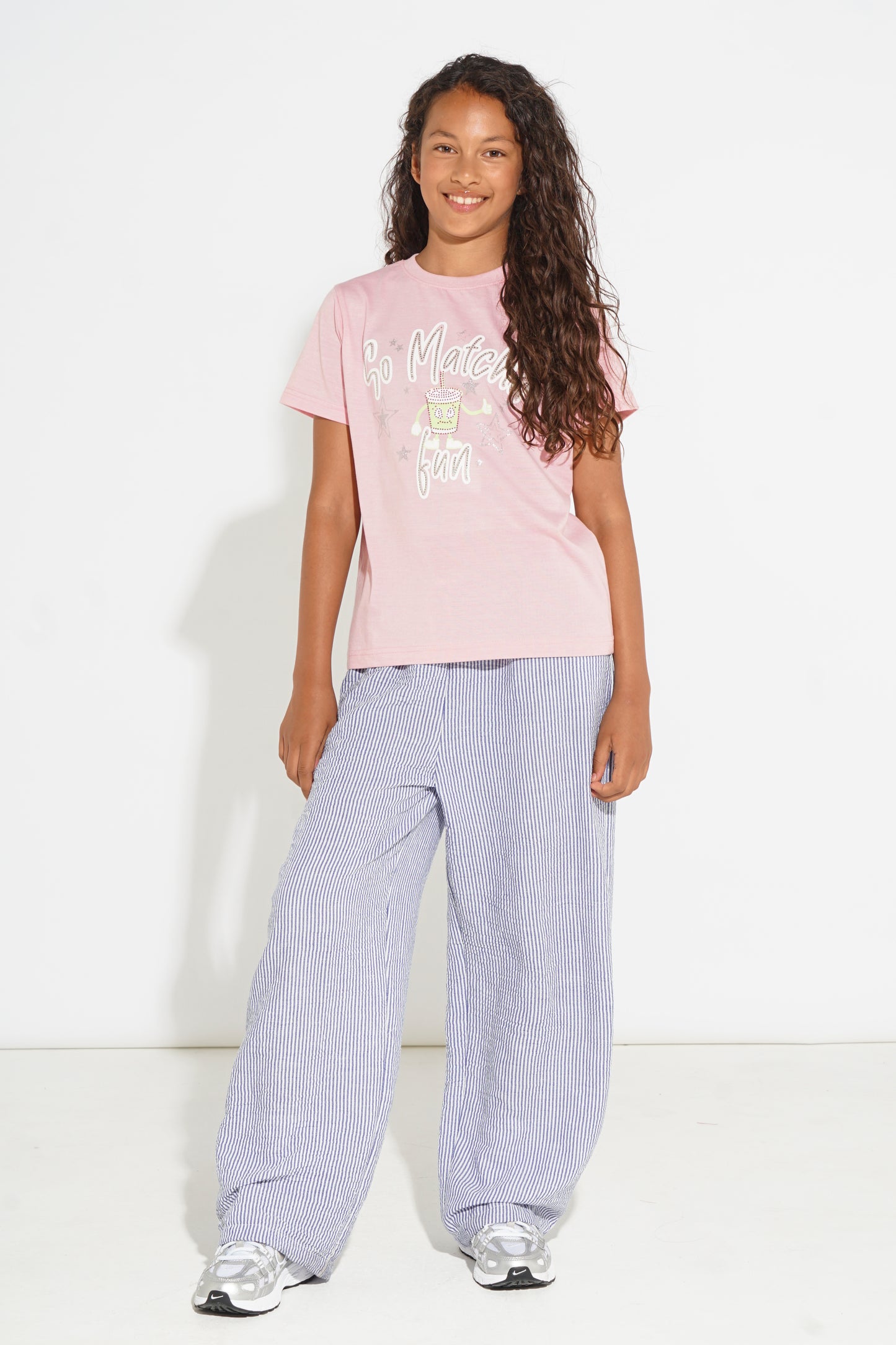 A young girl with long curly brown hair stands against a white studio background wearing a light pink graphic t-shirt, loose light purple wide-leg trousers, and silver sneakers.