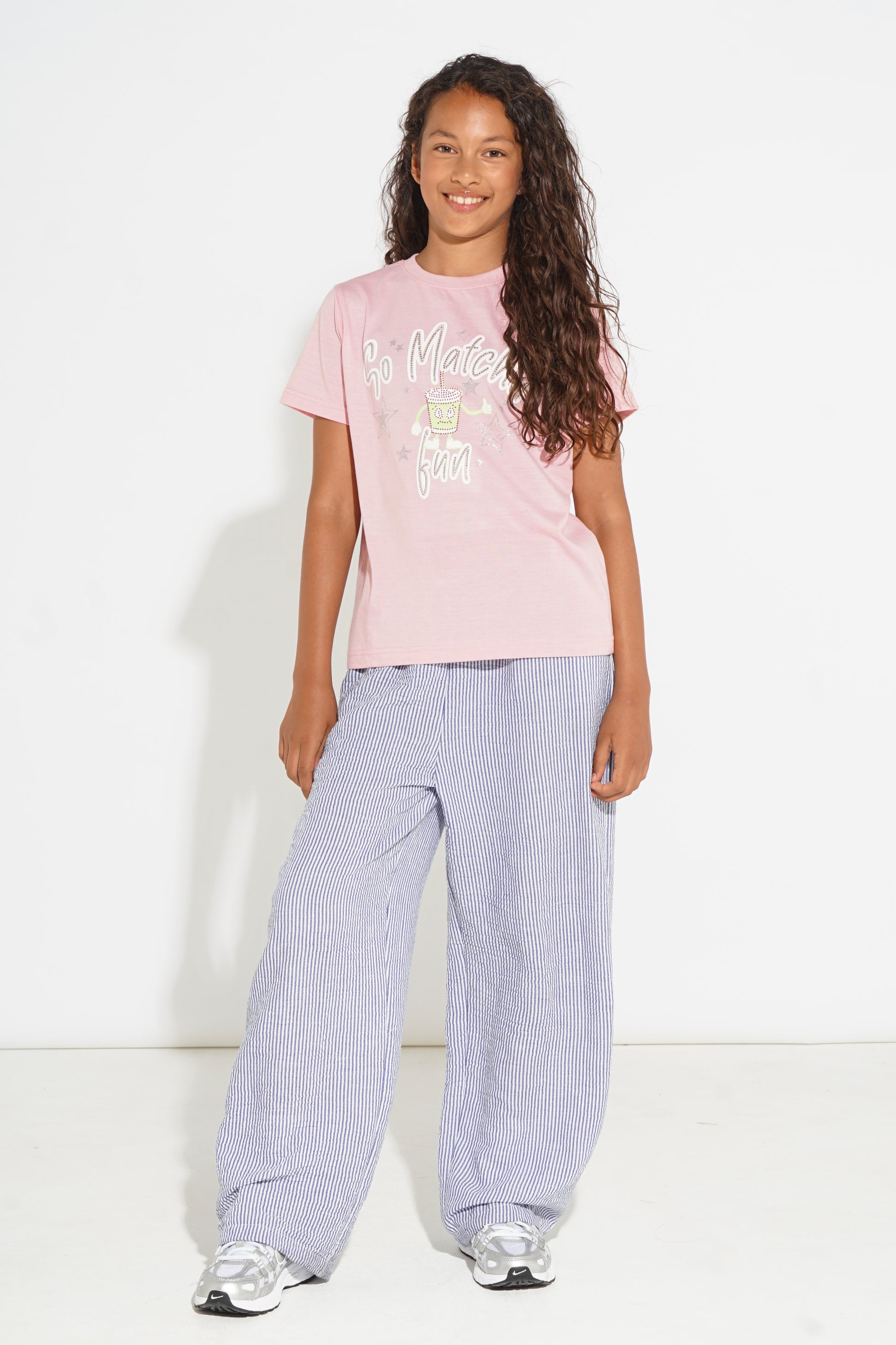 A young girl with long curly brown hair stands against a white studio background wearing a light pink graphic t-shirt, loose light purple wide-leg trousers, and silver sneakers.