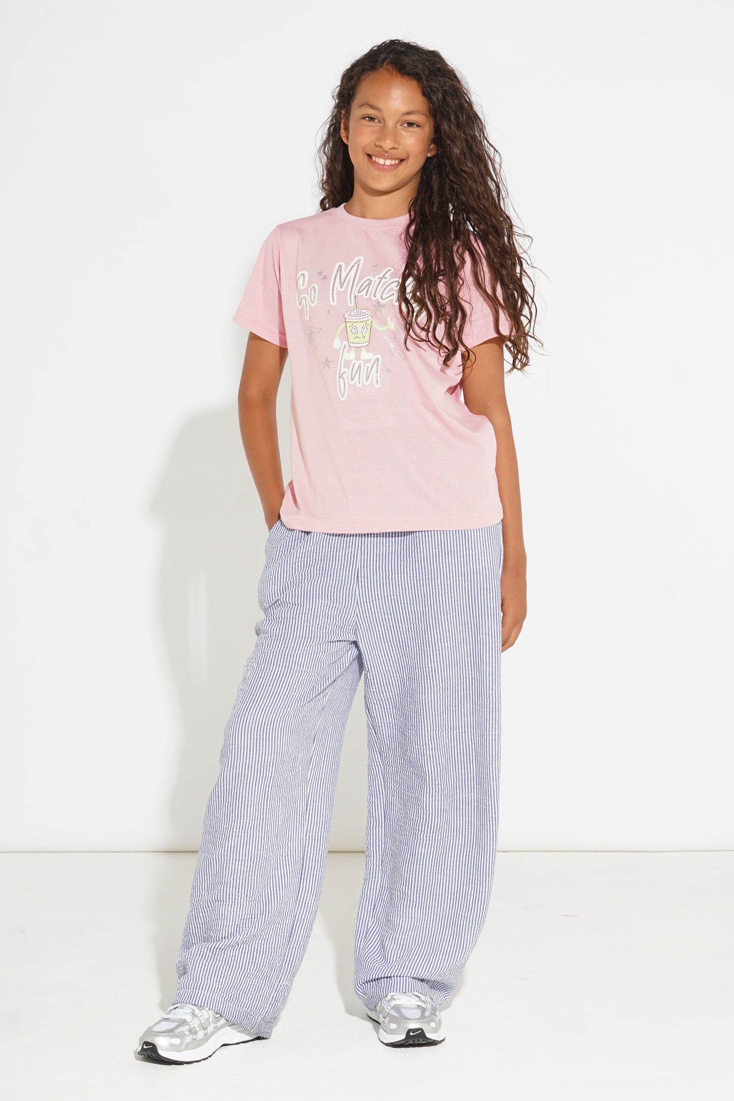 A young girl with long curly brown hair stands against a white studio background wearing a light pink graphic t-shirt, wide-leg light purple pants, and white sneakers.