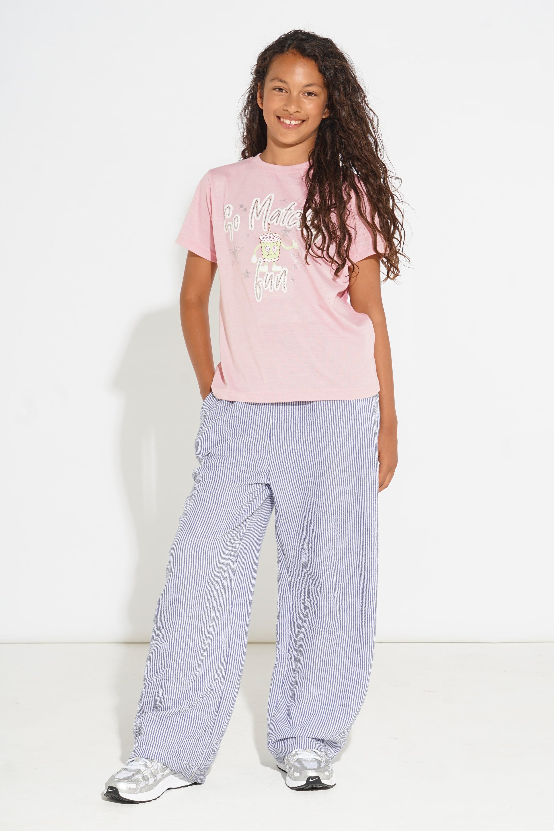 A young girl with long curly brown hair stands against a white studio background wearing a light pink graphic t-shirt, wide-leg light purple pants, and white sneakers.