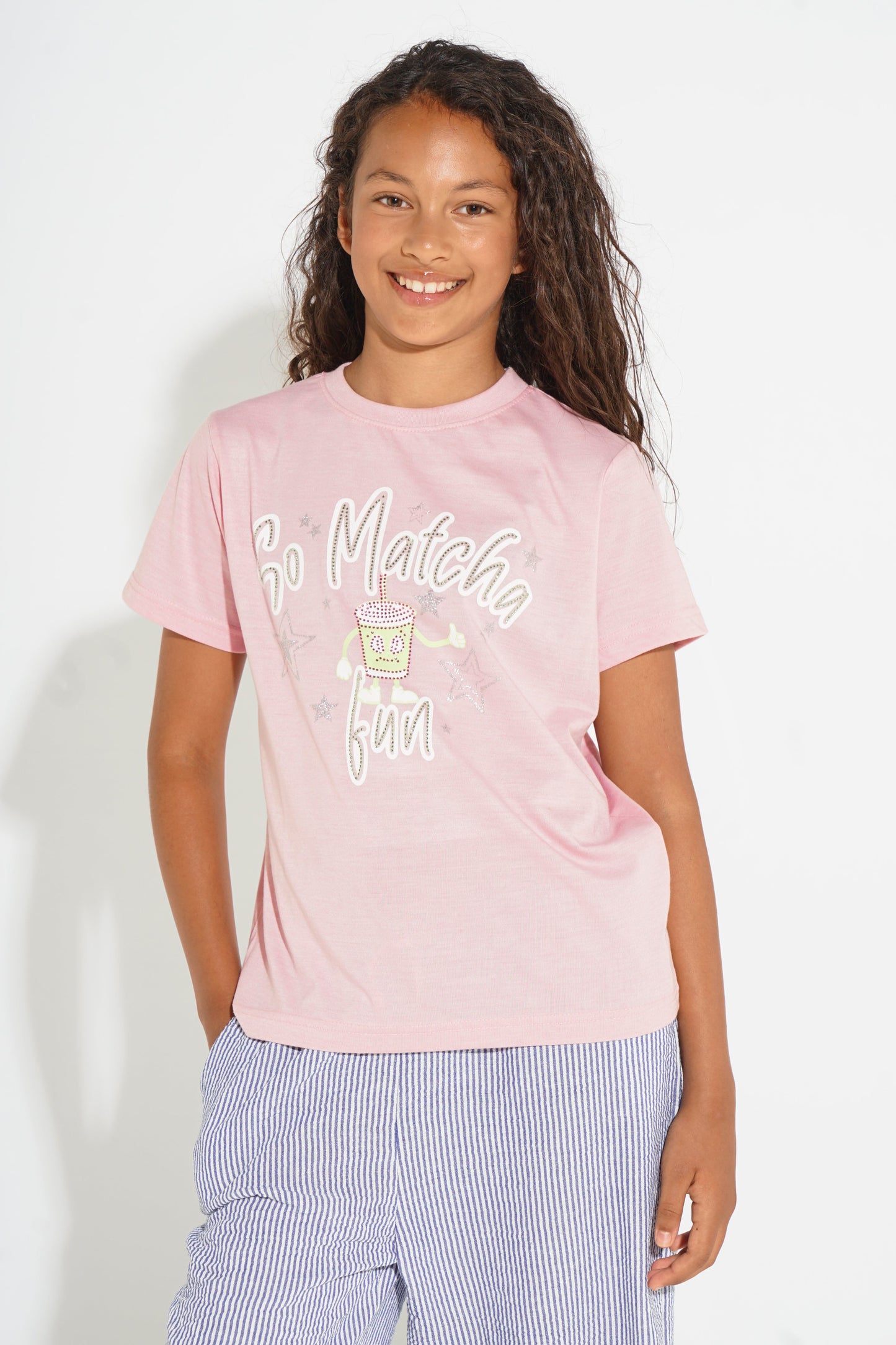 A young girl with long curly brown hair smiling, wearing a light pink graphic t-shirt with 'So Matcha Fun' written in white script, paired with light blue and white striped pants, against a plain white background.