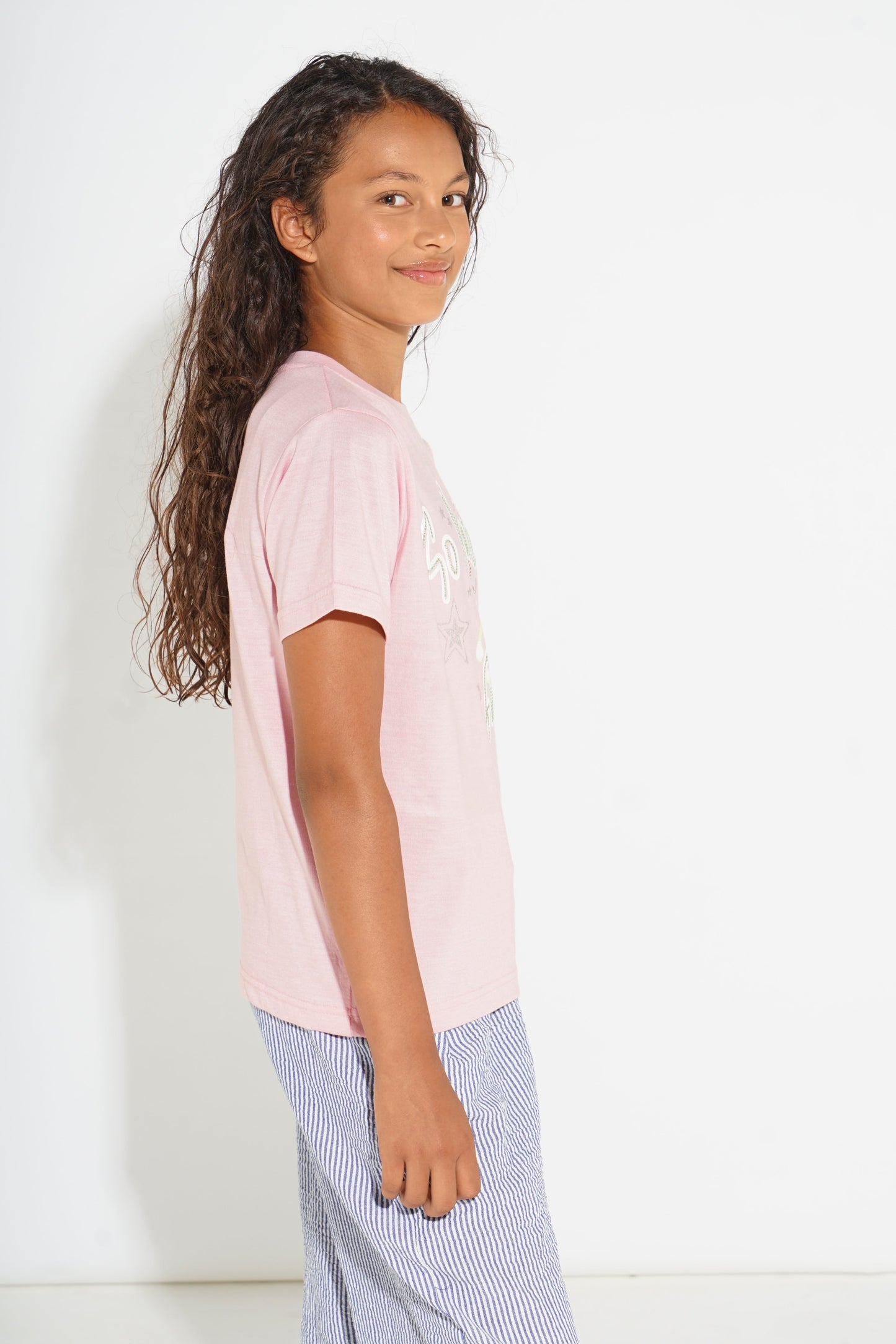 A young girl with long curly brown hair is shown from the side, wearing a light pink short-sleeve t-shirt with a white graphic print on the front and blue-and-white vertically striped pants, set against a plain white studio background.