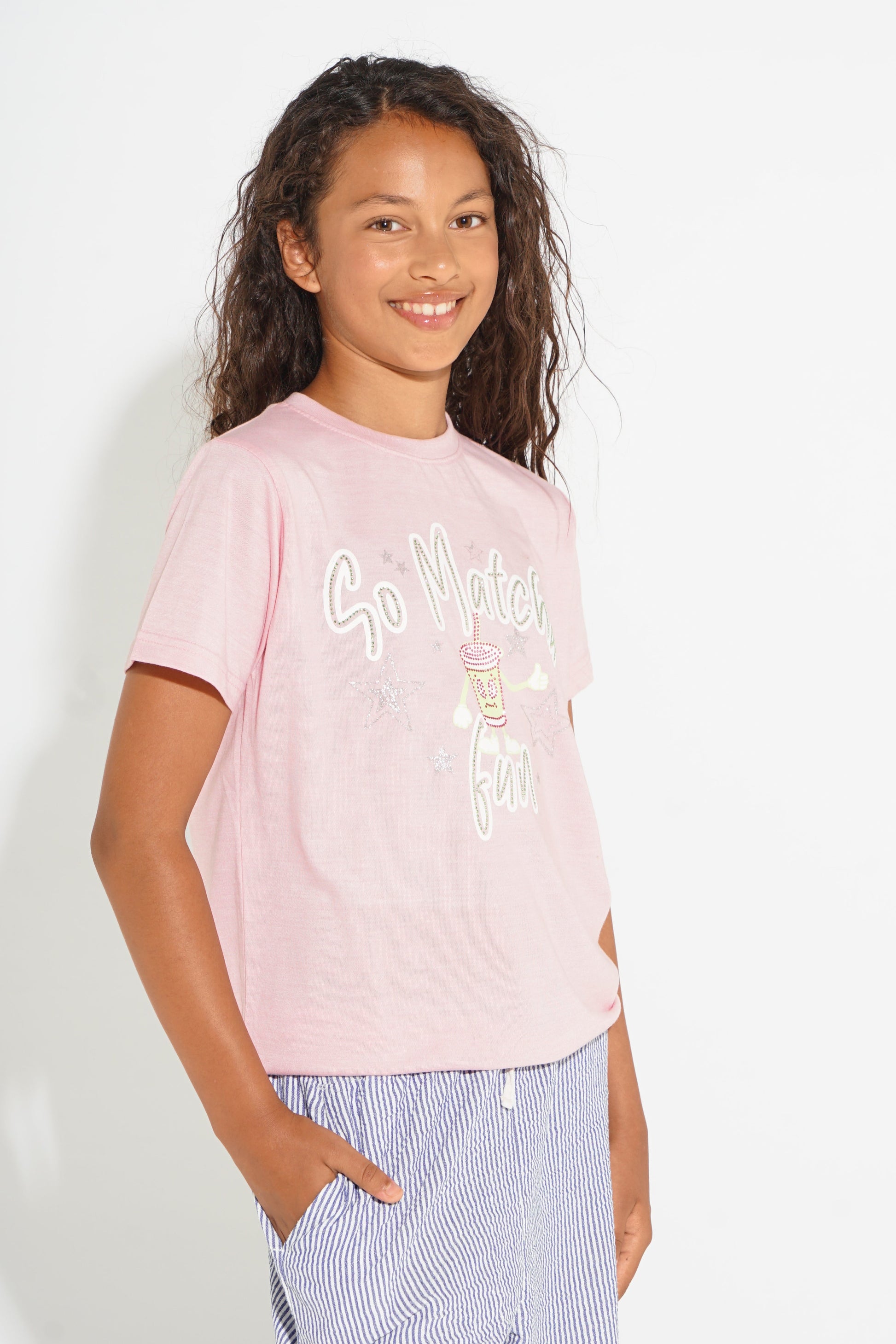A young girl with curly brown hair smiling, wearing a light pink graphic t-shirt with 'So Mooty Fun' printed on the front, paired with blue and white striped shorts, against a plain white background.