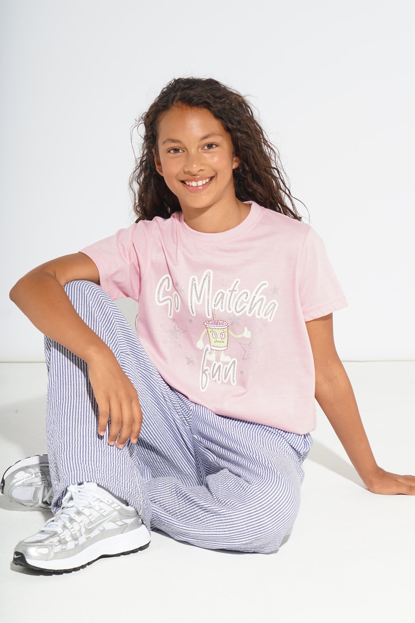 A young girl with curly brown hair sits cross-legged on a white studio floor, wearing a light pink graphic T-shirt that reads 'So Matcha Fun' with a matcha illustration, paired with light blue and white striped pants and white sneakers.