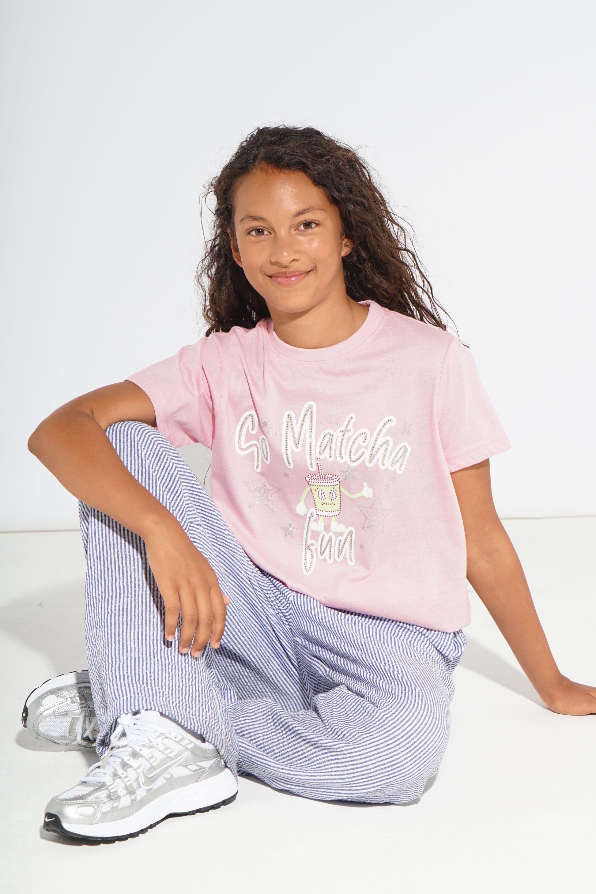 A young girl with curly brown hair sits on a white floor against a white wall, wearing a light pink graphic t-shirt that reads 'G. Matcha Fun' in white script, paired with blue and white striped pants and white sneakers.