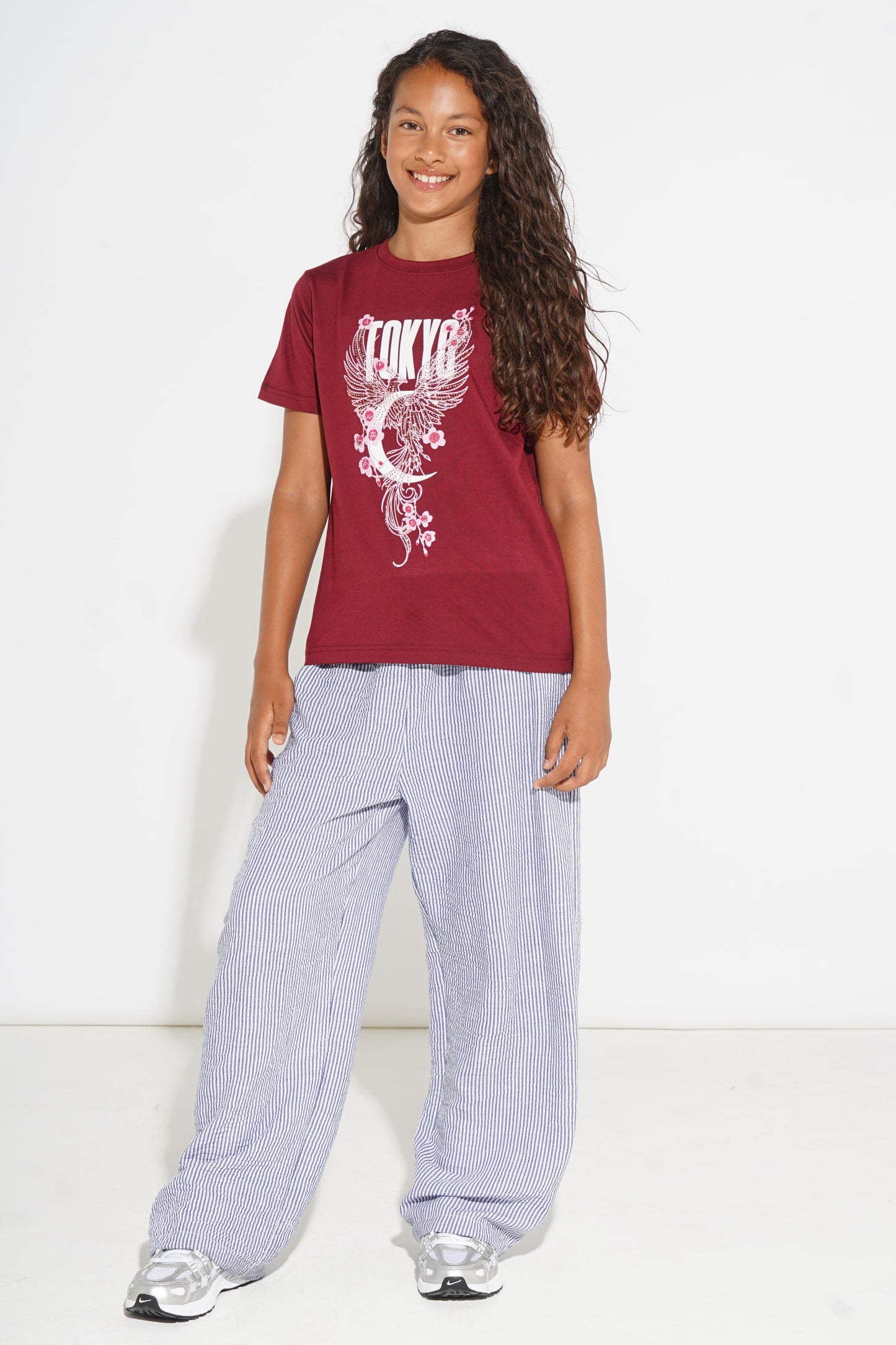 A young girl with long curly brown hair stands against a white studio background wearing a maroon short-sleeve graphic T-shirt with a white 'Tokyo' dragon print, loose light purple wide-leg pants, and silver sneakers.